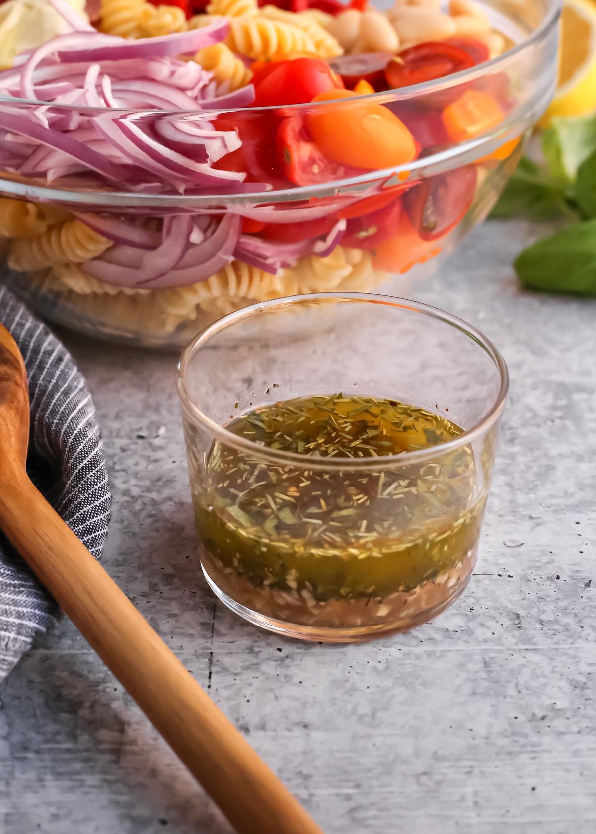 A small clear glass ramekin containing a homemade Italian-style vinaigrette on a kitchen countertop with a pasta salad and fresh basil in the background