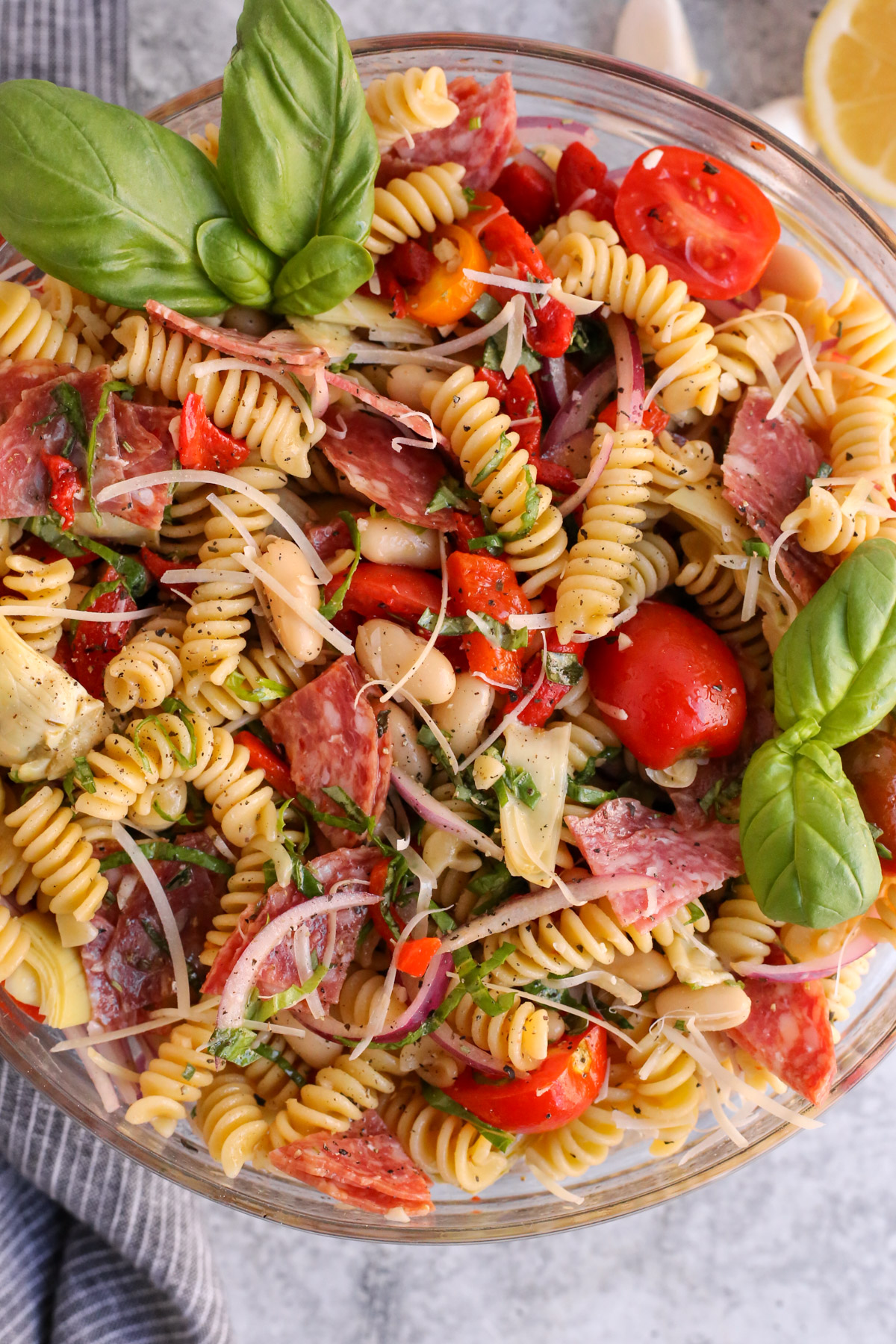 A closely cropped overhead view of antipasto pasta salad served in a clear glass mixing bowl and garnished with fresh basil, shredded parmesan cheese, and Italian herbs