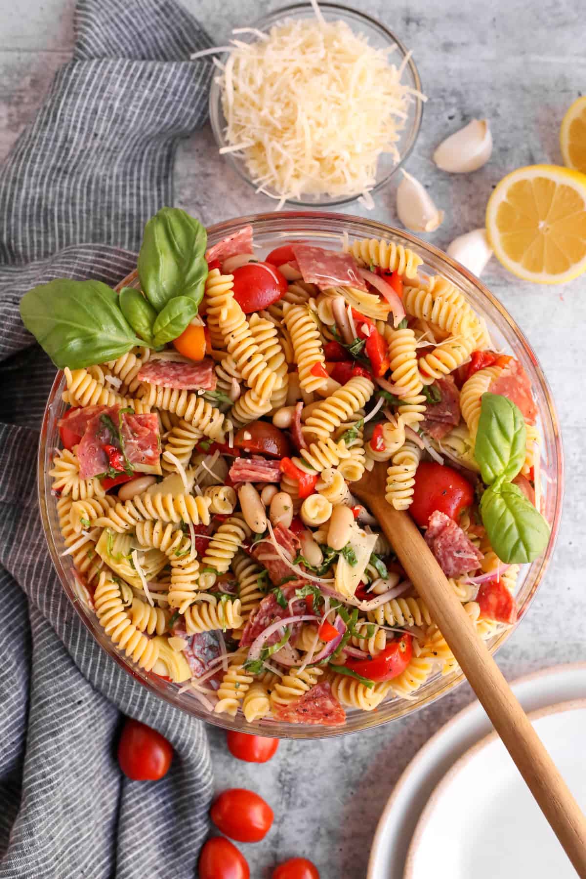 An overhead view of a clear glass mixing bowl containing a prepared antipasto pasta salad, with fresh basil for garnish and extra lemon wedges and parmesan cheese served with it