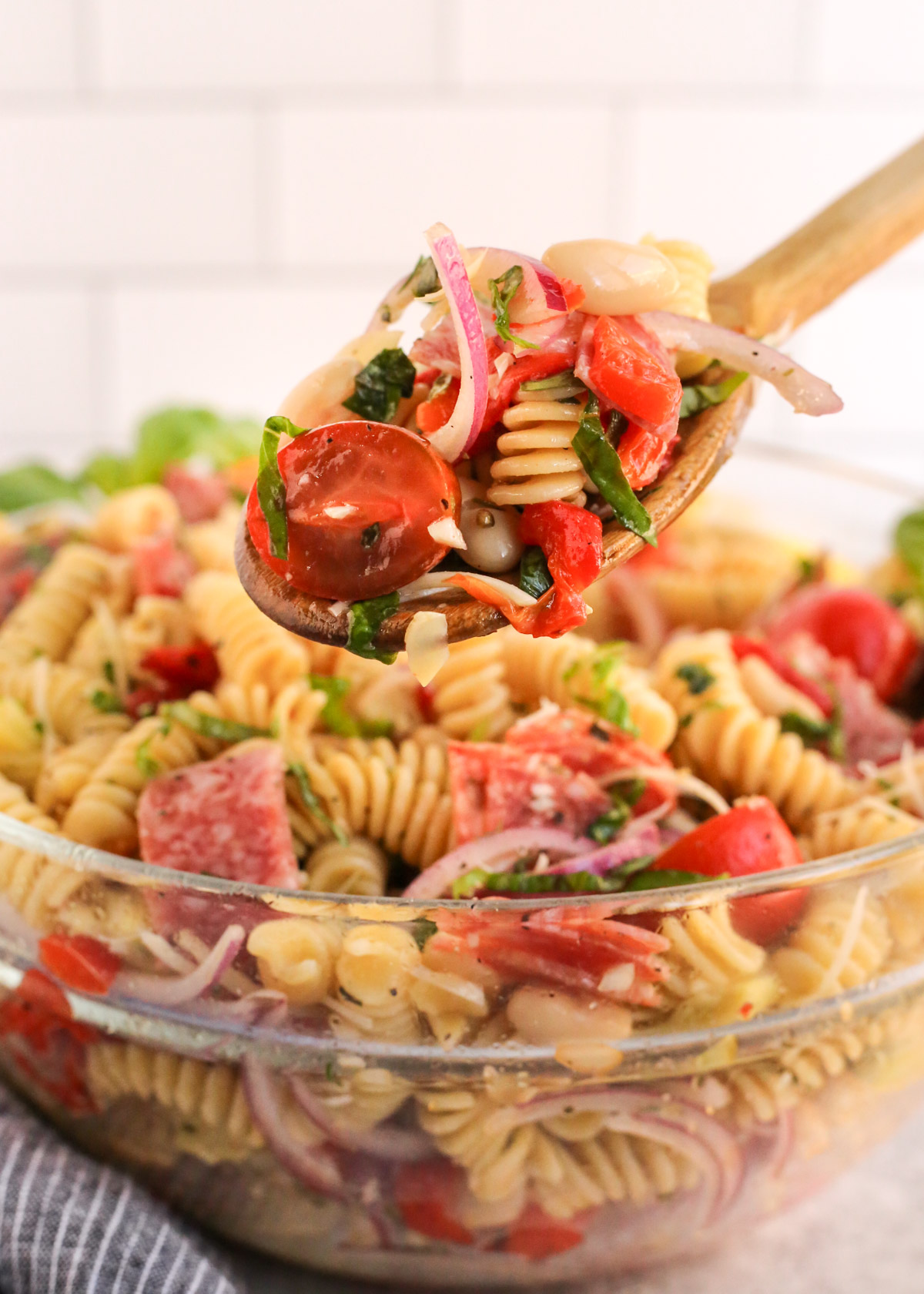 A wooden serving spoon lifts a spoonful of antipasto pasta salad out of the mixing bowl, showing a variety of ingredients like rotini pasta, cherry tomatoes, sliced red onions, chopped basil, and pepperoni