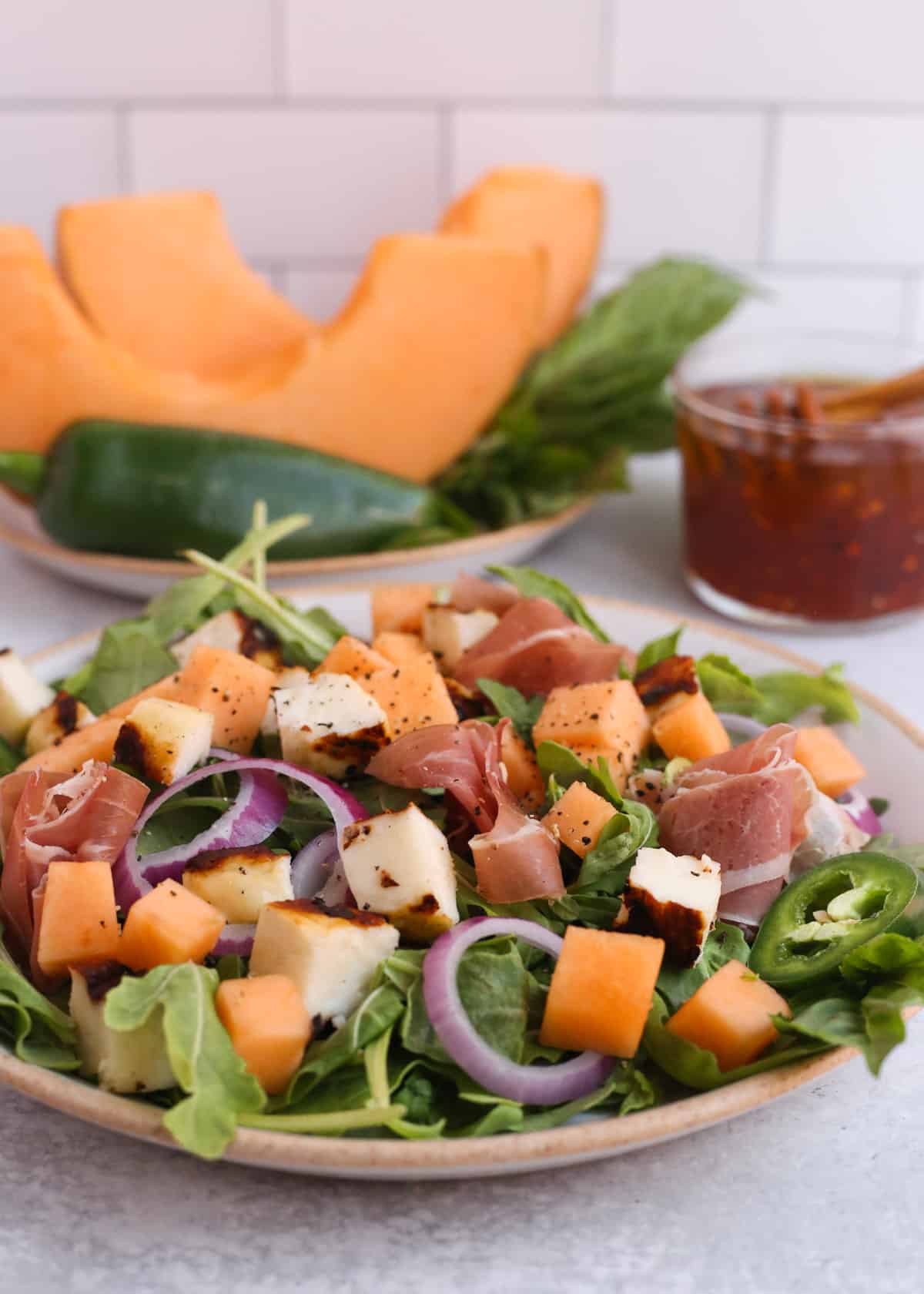 A ceramic plate filled with halloumi salad and garnished with cantaloupe, hot honey dressing, red onion, and prosciutto is served on a kitchen countertop with additional ingredients in the background