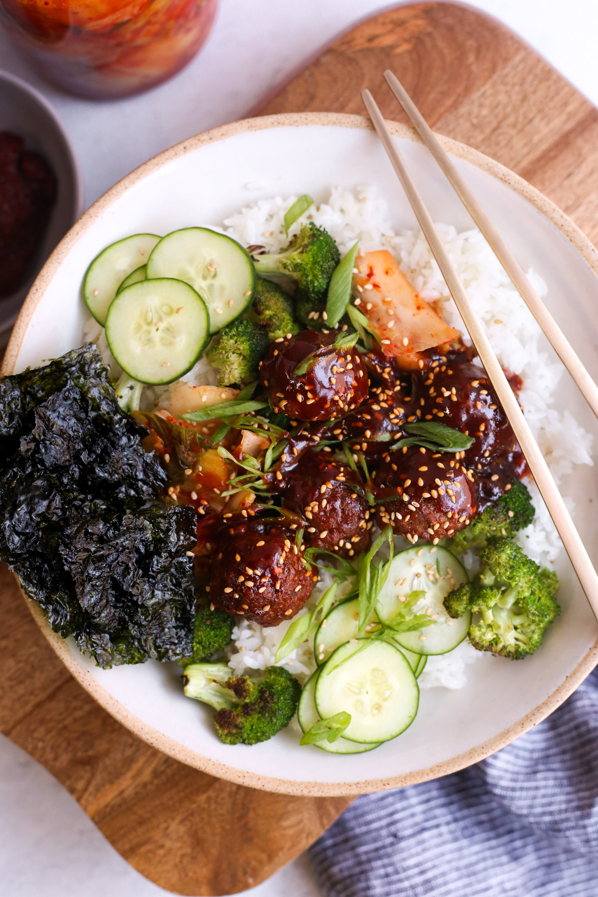 A rice bowl containing white rice, gochujang meatballs, sliced cucumbers, roasted broccoli, kimchi, and nori, garnished with toasted sesame seeds