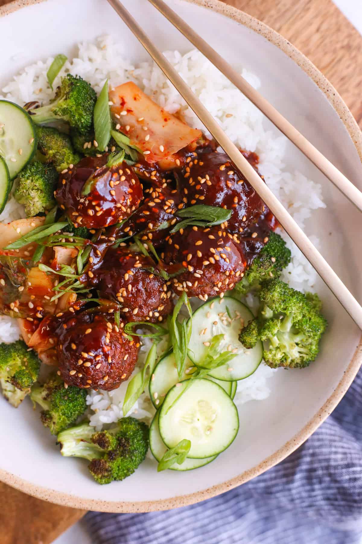 A top view of a bowl full of Korean-inspired gochujang meatballs, garnished with kimchi, sliced cucumbers, roasted broccoli, and served over white rice