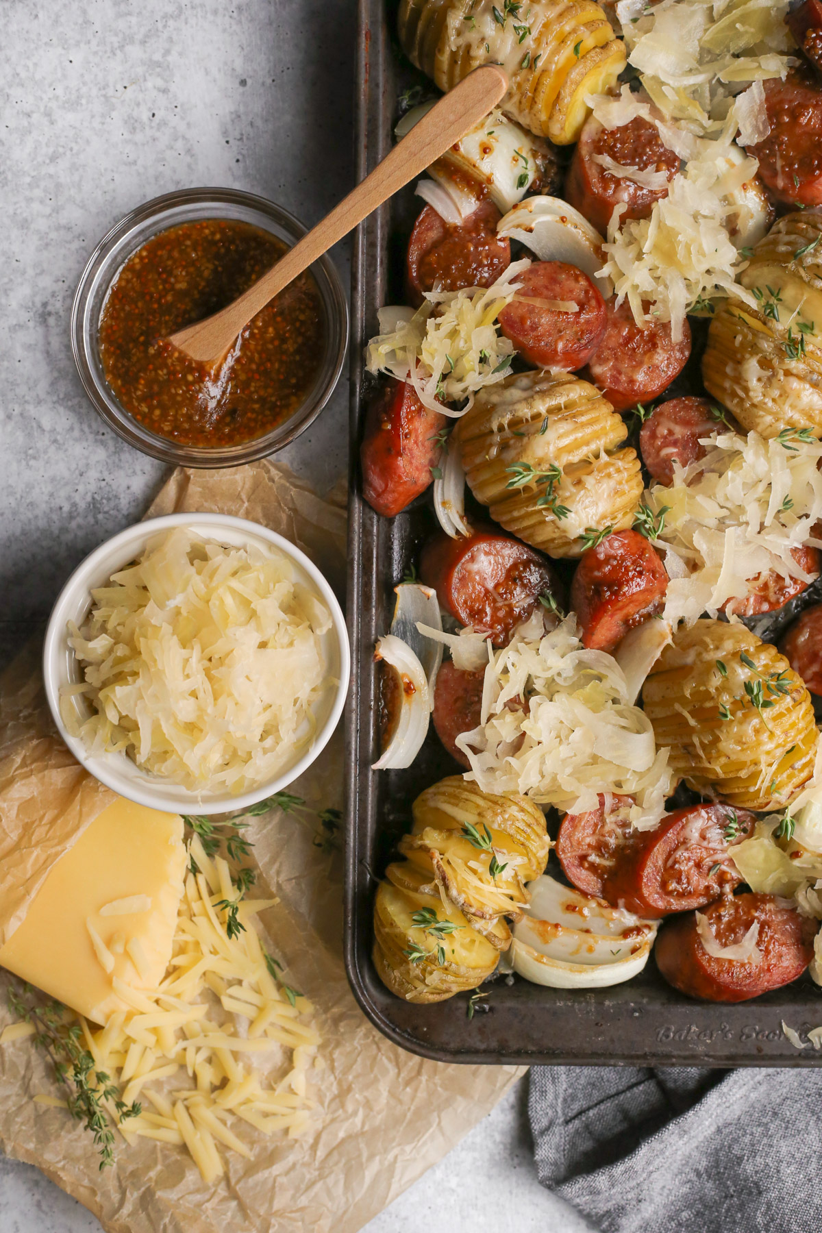 A cropped overhead view of a kielbasa sheet pan dinner recipe, served on a well seasoned sheet pan with dark patina, with cheesy hasselback potatoes, sauerkraut, and roasted onions also displayed on the pan