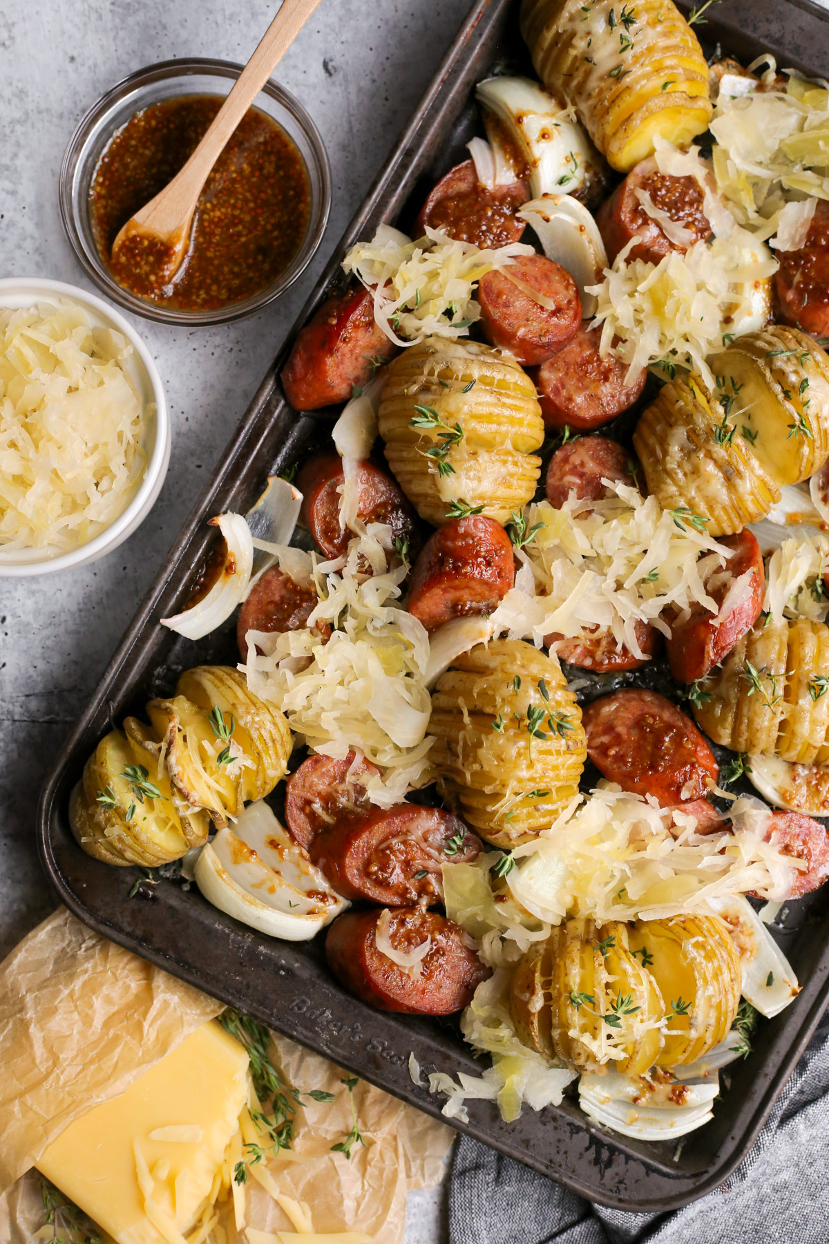 An overhead view of a kielbasa sheet pan dinner recipe with cheesy hasselback potatoes, sliced onions, sauerkraut, and a sweet mustard glaze on a kitchen countertop with extra ingredients surrounding it