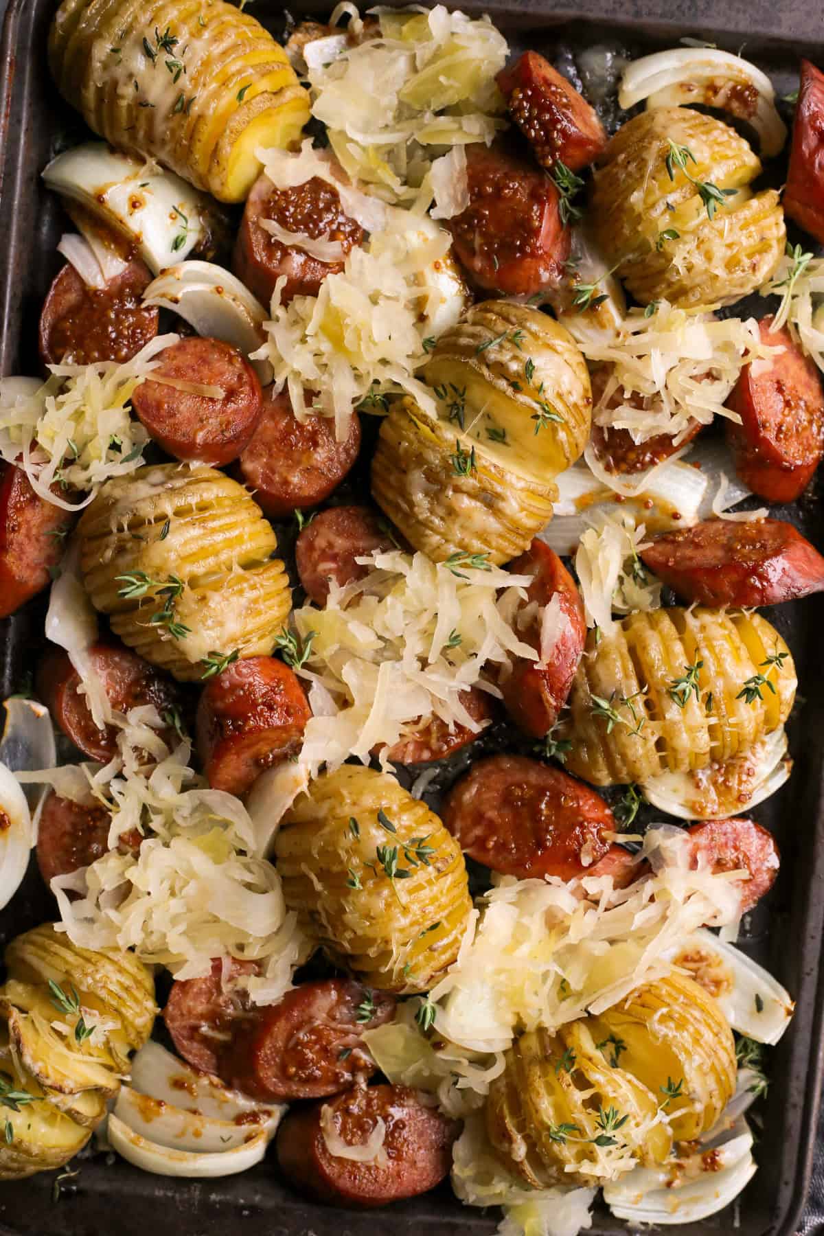 A very closely cropped overhead view of sliced kielbasa sausage, cheesy hasselback potatoes, sliced onions, and sauerkraut on a dark metal sheet pan, garnished with fresh thyme