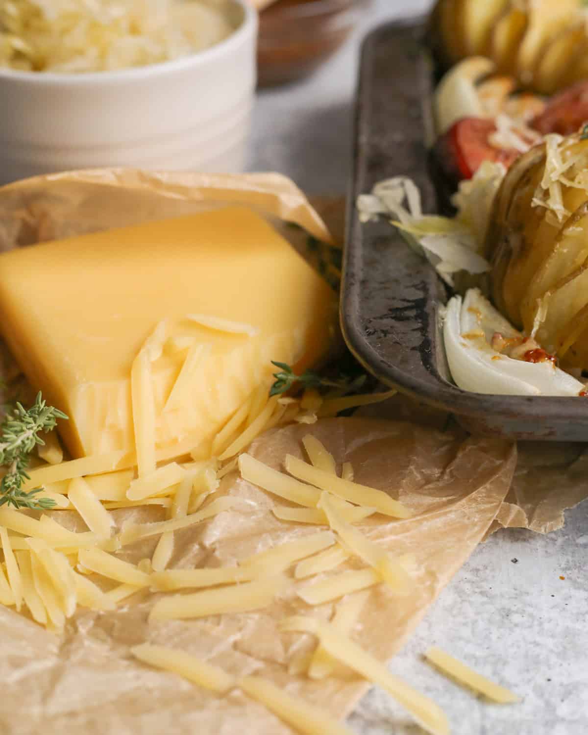 A block of cheese sits on a wrinkled piece of parchment paper, partially grated, with small clumps of shredded cheese arranged around it. The corner of a dark metal sheet pan is visible on the right side and other ingredients are positioned in the background