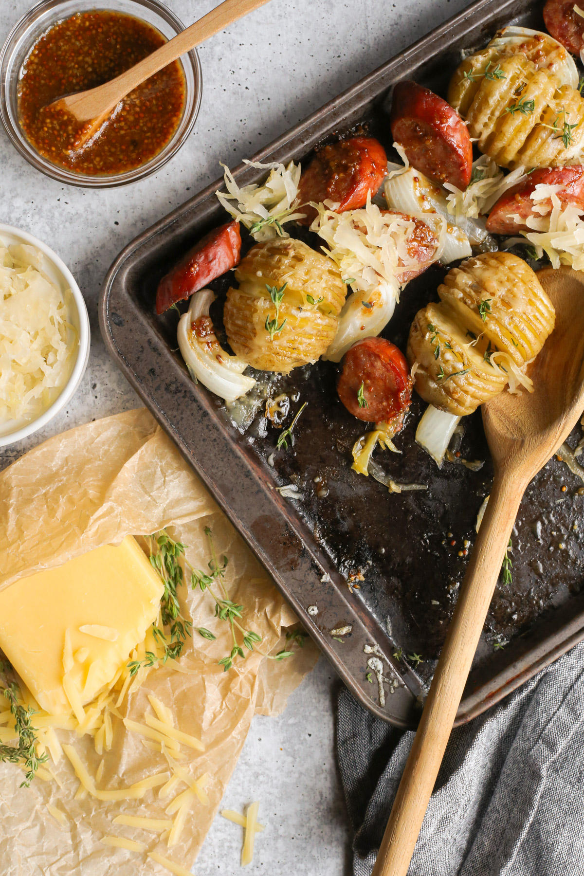 A messy overhead view of a kielbasas sheet pan dinner, with a wooden serving spoon and small dishes with a sweet mustard glaze, shredded cheese, and fresh thyme