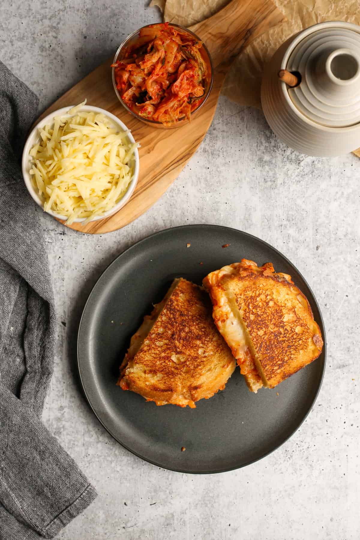 Overhead view of a kimchi grilled cheese sandwich served on a black ceramic plate, accompanied with extra kimchi and shredded cheese
