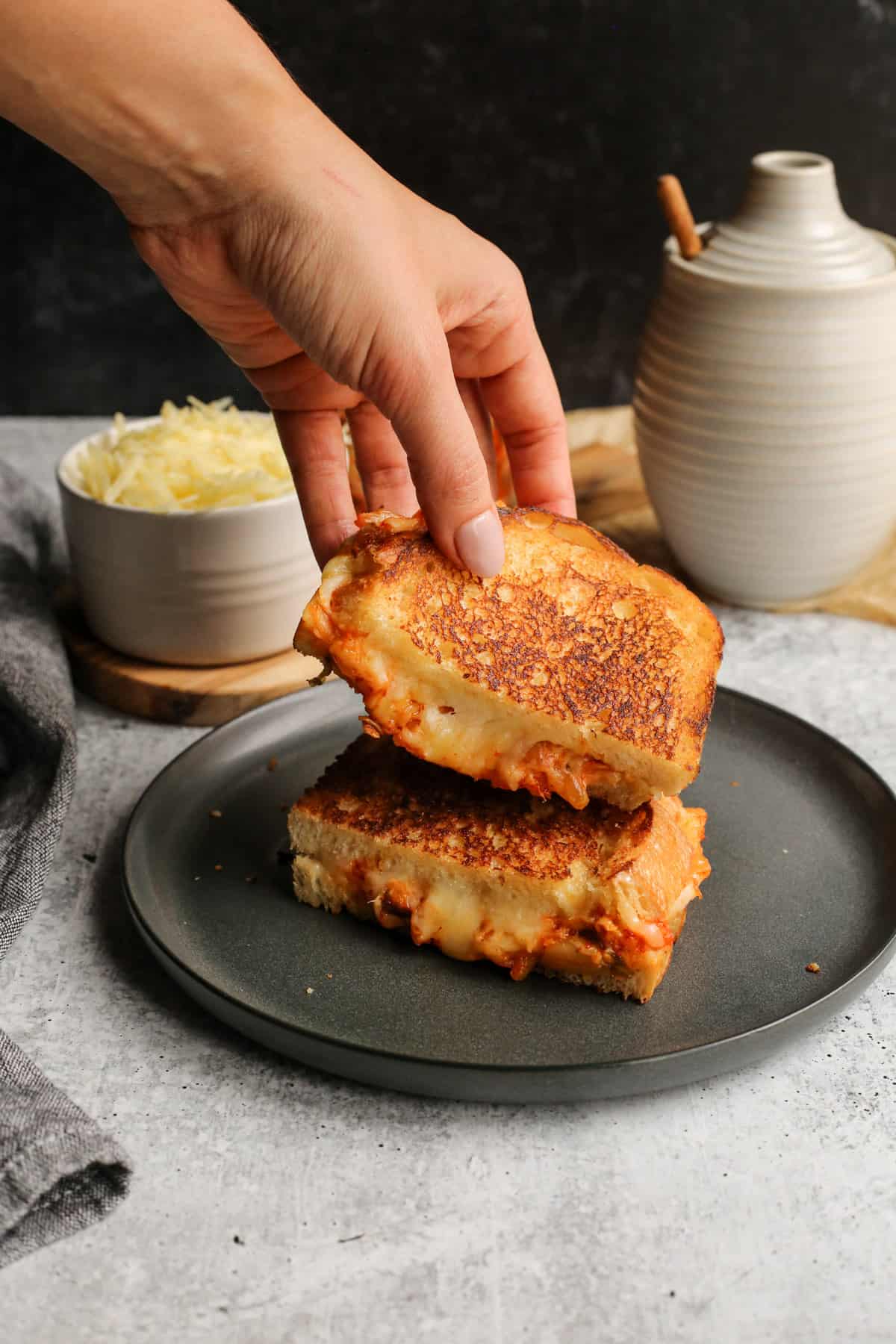 A woman's hand reaches into the frame to pick up half of a sandwich. You can see melted cheese and kimchi inside the toasted bread