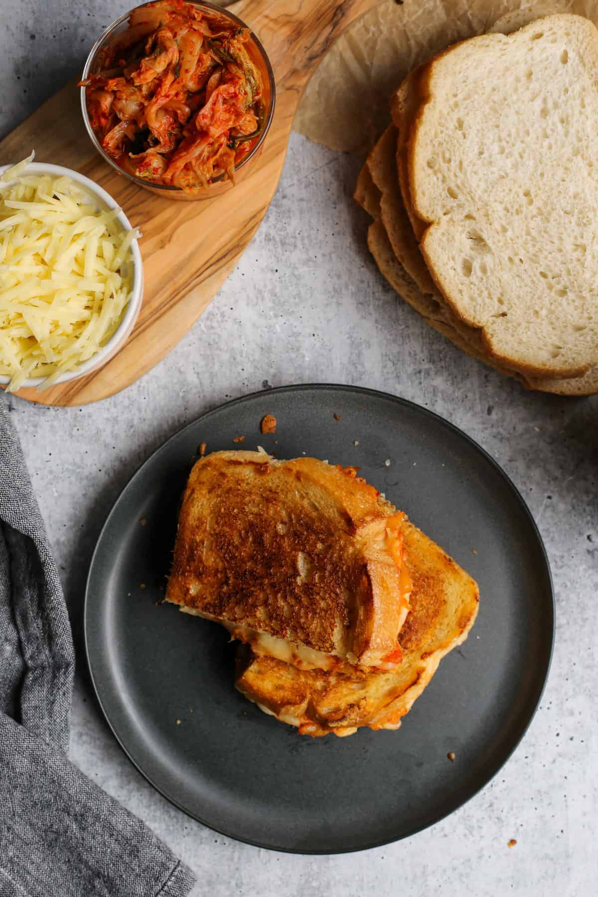 Overhead view of a kimchi grilled cheese sandwich on a black ceramic dishes, surrounded by additional ingredients to make more sandwiches