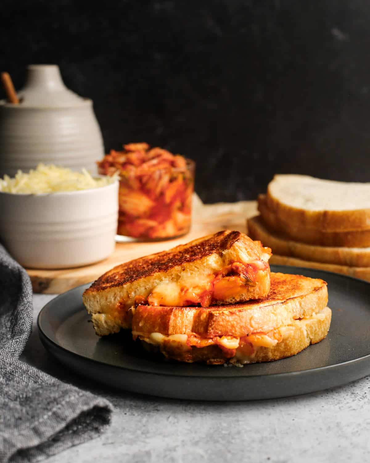 Side view of a kimchi grilled cheese sandwich served on a black ceramic plate. In the background are ramekins filled with extra kimchi and shredded cheese and slices of sourdough bread