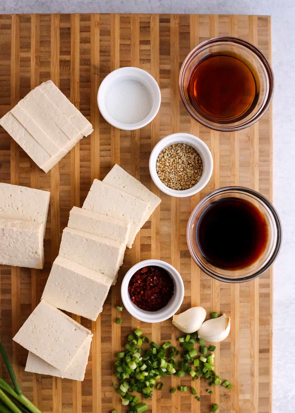 Overhead view of a block of tofu, sliced into squares, on a wooden butcher block cutting board surrounded by the rest of the ingredients needed to make Korean braised tofu, or dubu jorim.