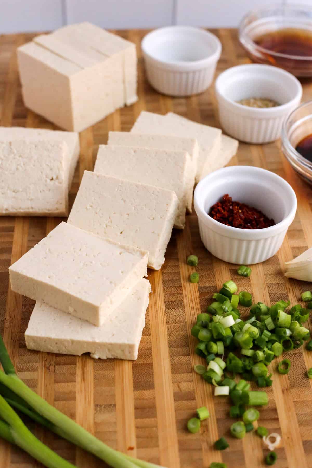 A wooden butcher block cutting board with a block of firm tofu, drained and pressed dry and cut into squares with sliced green onions and variously other in