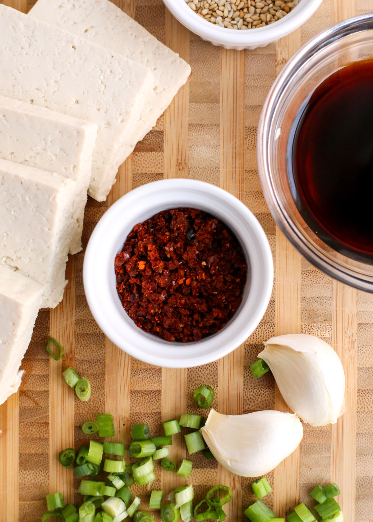 Overhead view of a small ramekin of gochugaru, Korean red chili pepper flakes, displayed on a wooden cutting board with sliced firm tofu, sliced green onions, garlic cloves, and sauce ingredients of dubu jorim