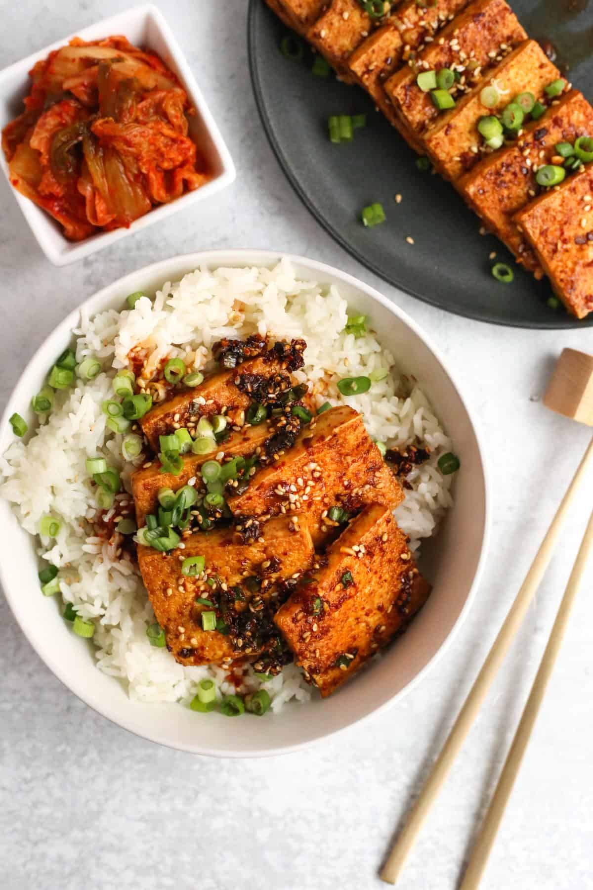 Overhead view of a white ceramic bowl filled with white rice, topped with pieces of dubu jorim (Korean braised tofu) along with toasted sesame seeds, sliced green onions, and extra sauce. A pair of brass metal chopsticks is placed next to the bowl and a side dish of kimchi and the remaining dubu jorim are shown next to the serving bowl