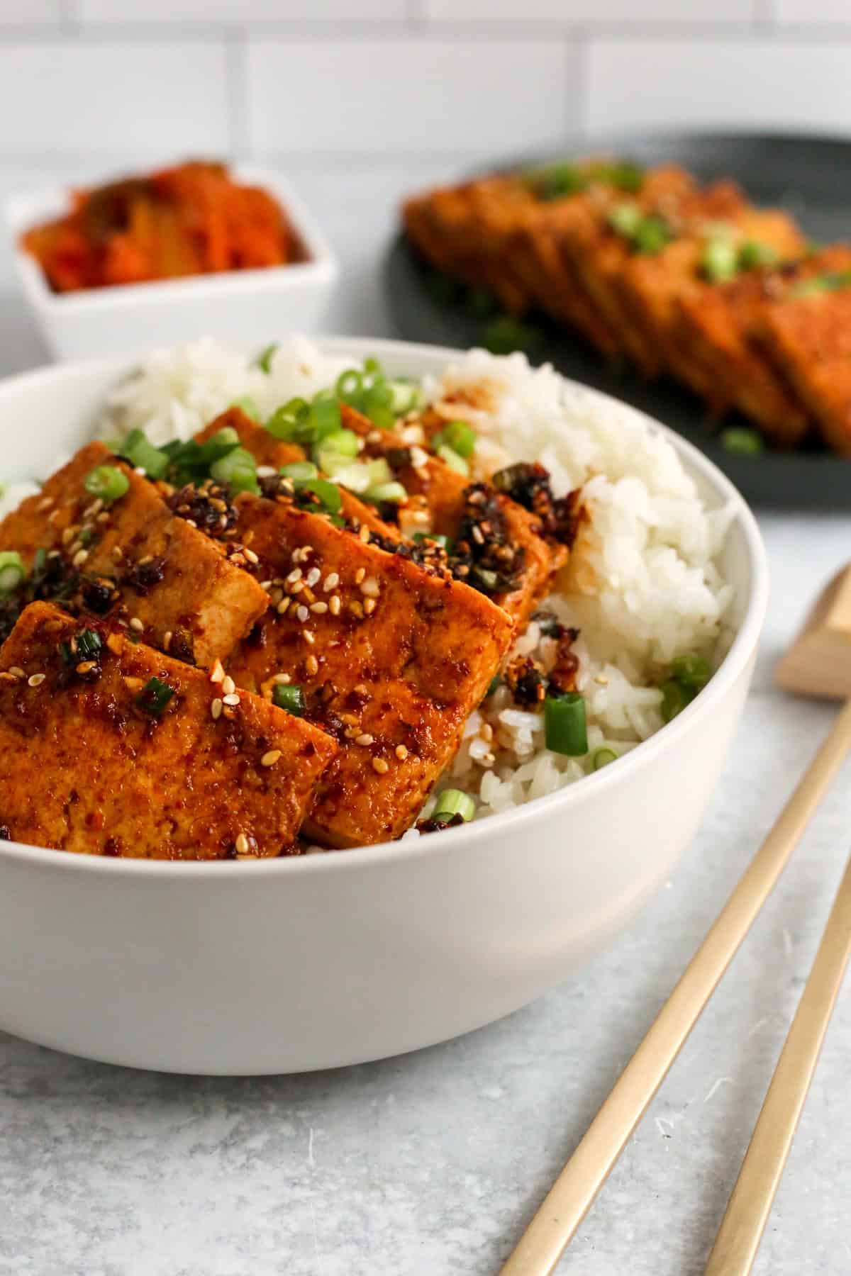 A white ceramic bowl filled with white rice is topped with pieces of dubu jorim, Korean braised tofu, along with toasted sesame seeds, sliced green onions, and extra sauce. A pair of brass metal chopsticks is placed next to the bowl