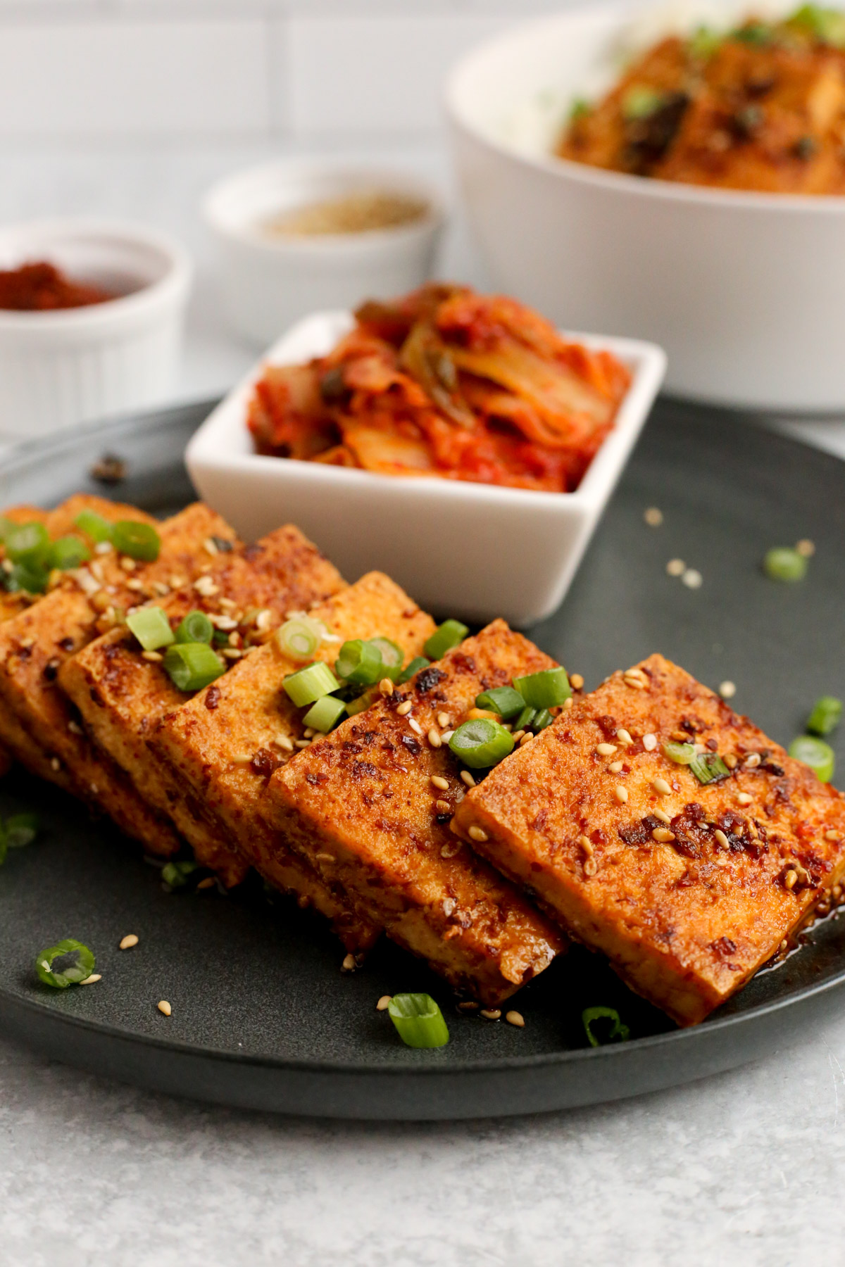 A close shot of dubu jorim, Korean braised tofu, stacked and served on a dark ceramic dish with kimchi, garnishes, and a rice bowl visible in the background