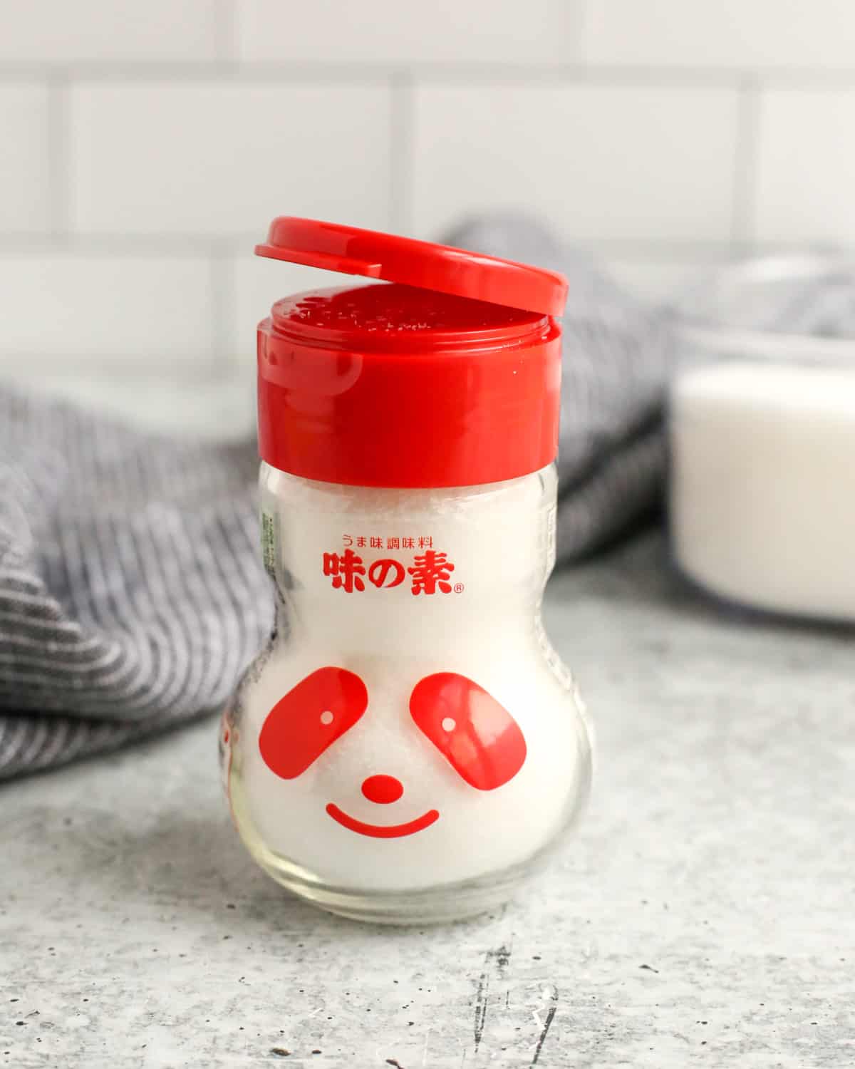 A shaker of MSG, which can be used to make low sodium foods taste better, on a countertop with the lid slightly popped open, with red markings that resemble the face of a panda