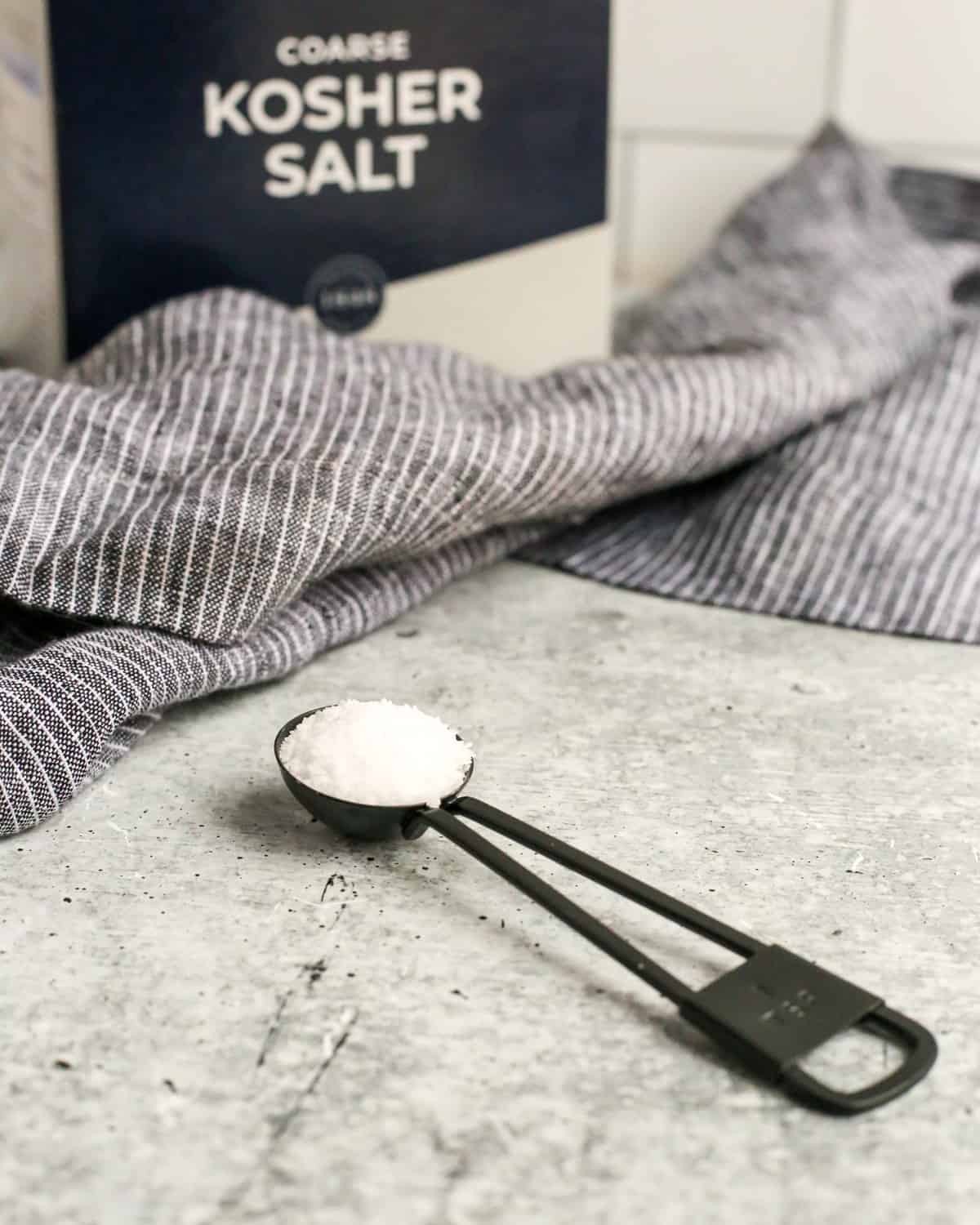 A black teaspoon full of coarse kosher salt rests on a cement colored kitchen countertop in front of a box of Morton's coarse Kosher salt with the labeling and letters visible in the background