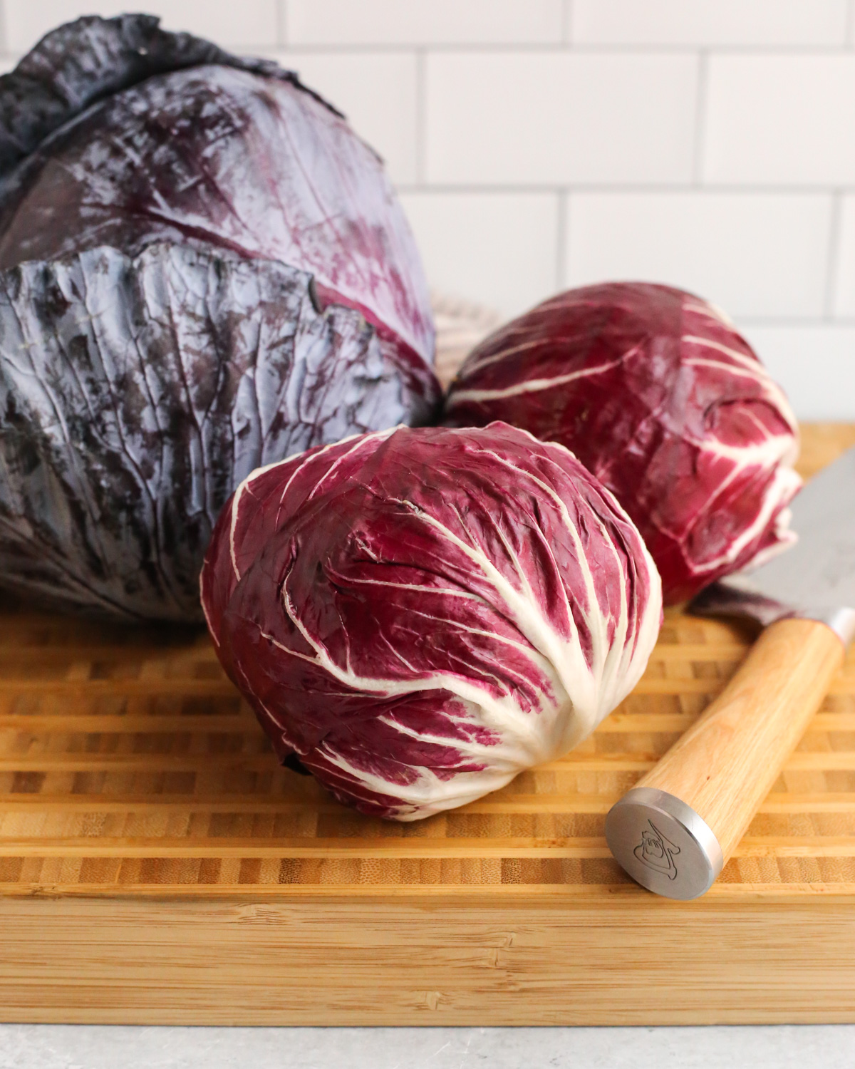 Brightly lit image of two heads of radicchio on a wooden butcher block cutting board, positioned in front of a large head of red cabbage