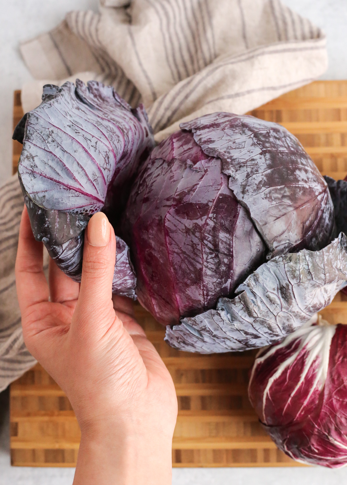 A woman's hand peels back the outer leaves on a head of red cabbage, indicating this is an important prep step prior to cutting, cooking, or serving