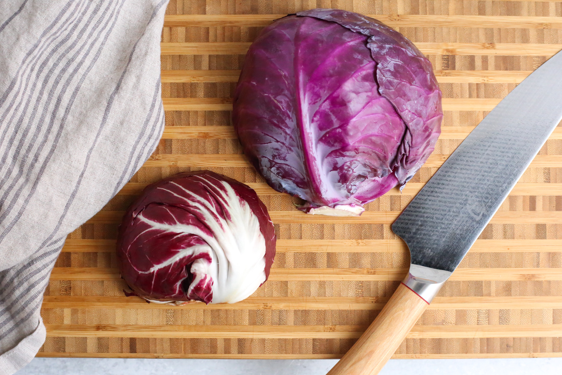 Overhead view of a butcher block cutting board with a head of radicchio next to a head of red cabbage, showing the subtle difference in size, color, and variegated white veins