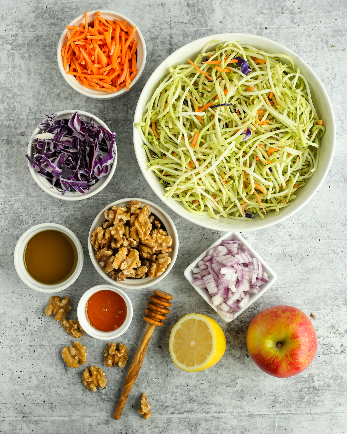 Flatlay on a kitchen countertop with the ingredients needed for this broccoli salad, including broccoli slaw, matchstick carrots, shredded red cabbage, diced red onions, walnuts, and the components of a dairy-free dressing