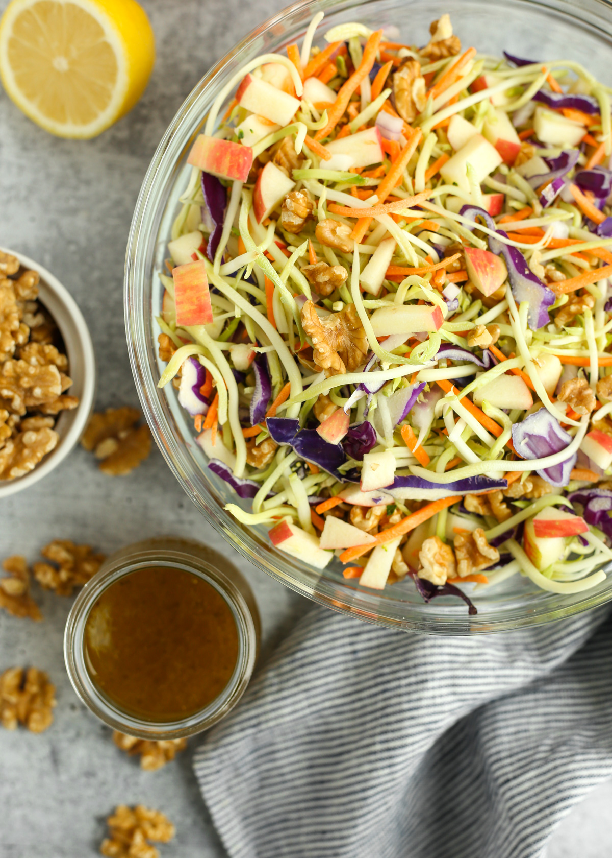 Overhead image of an undressed mixture of broccoli slaw, carrots, red cabbage, diced red onion and apples, and walnut pieces, with a dairy-free dressing on the side about to be mixed in