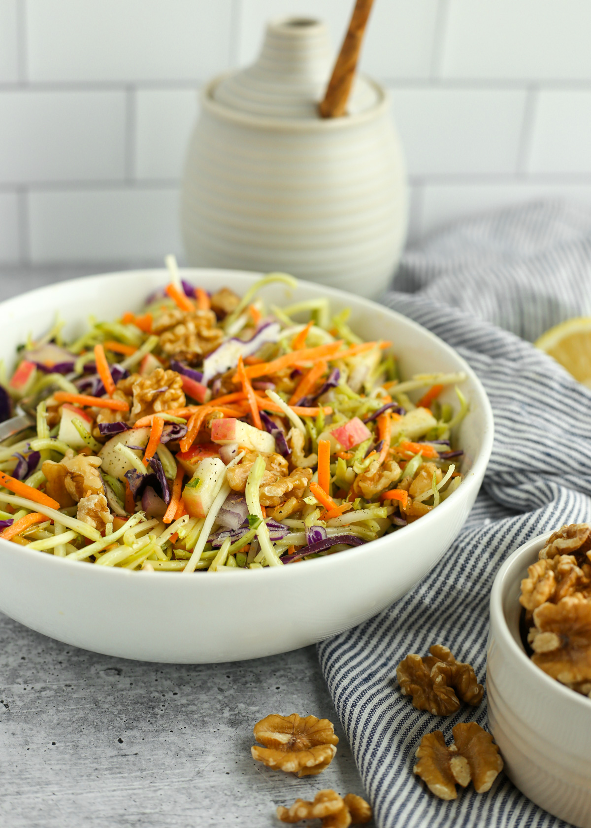 Angled view of a kitchen scene with a white serving bowl filled with a salad made from broccoli slaw, shredded carrots and red cabbage, walnuts, and diced apples, with additional ingredients and a blue and white striped dish towel arranged in the background