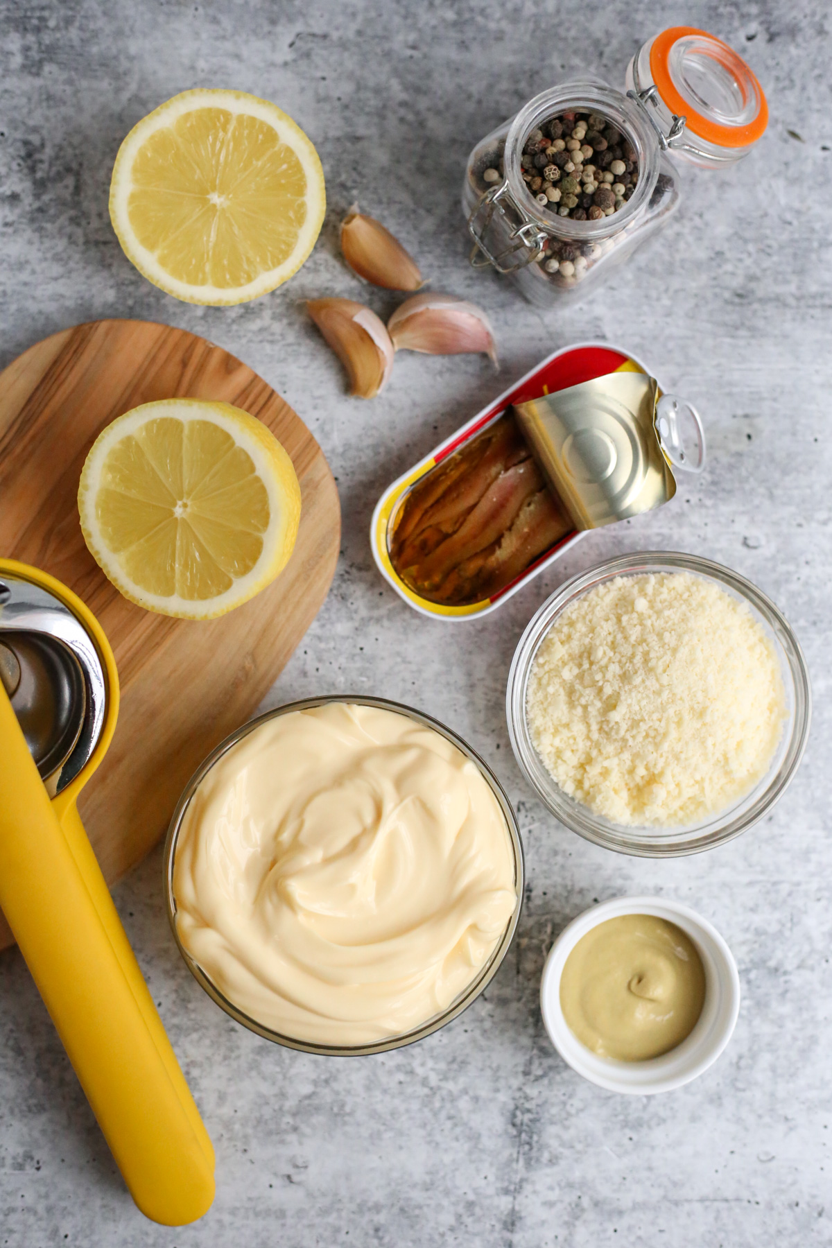 A flatlay of all the ingredients needed to make a homemade caesar salad dressing, including kewpie mayo, dijon mustard, anchovy fillets, black pepper, grated parmesan cheese, cloves of garlic, and a fresh lemon sliced in half