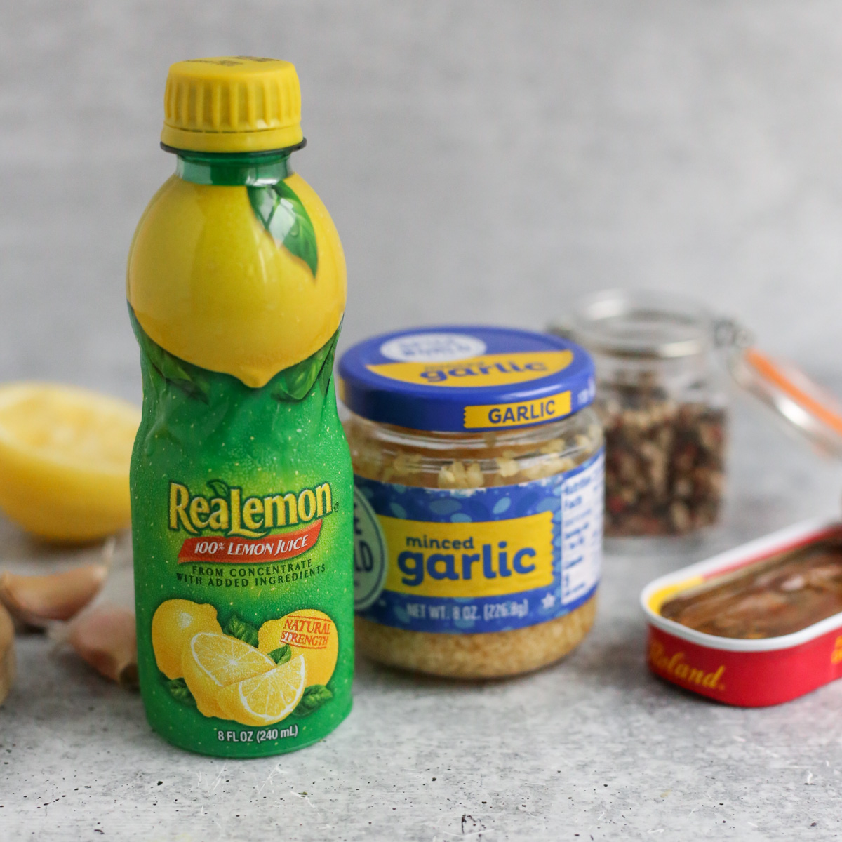 A bottle of lemon juice and jar of minced garlic are displayed on a kitchen countertop alongside garlic cloves, peppercorns, and a tin of anchovies packed in oil