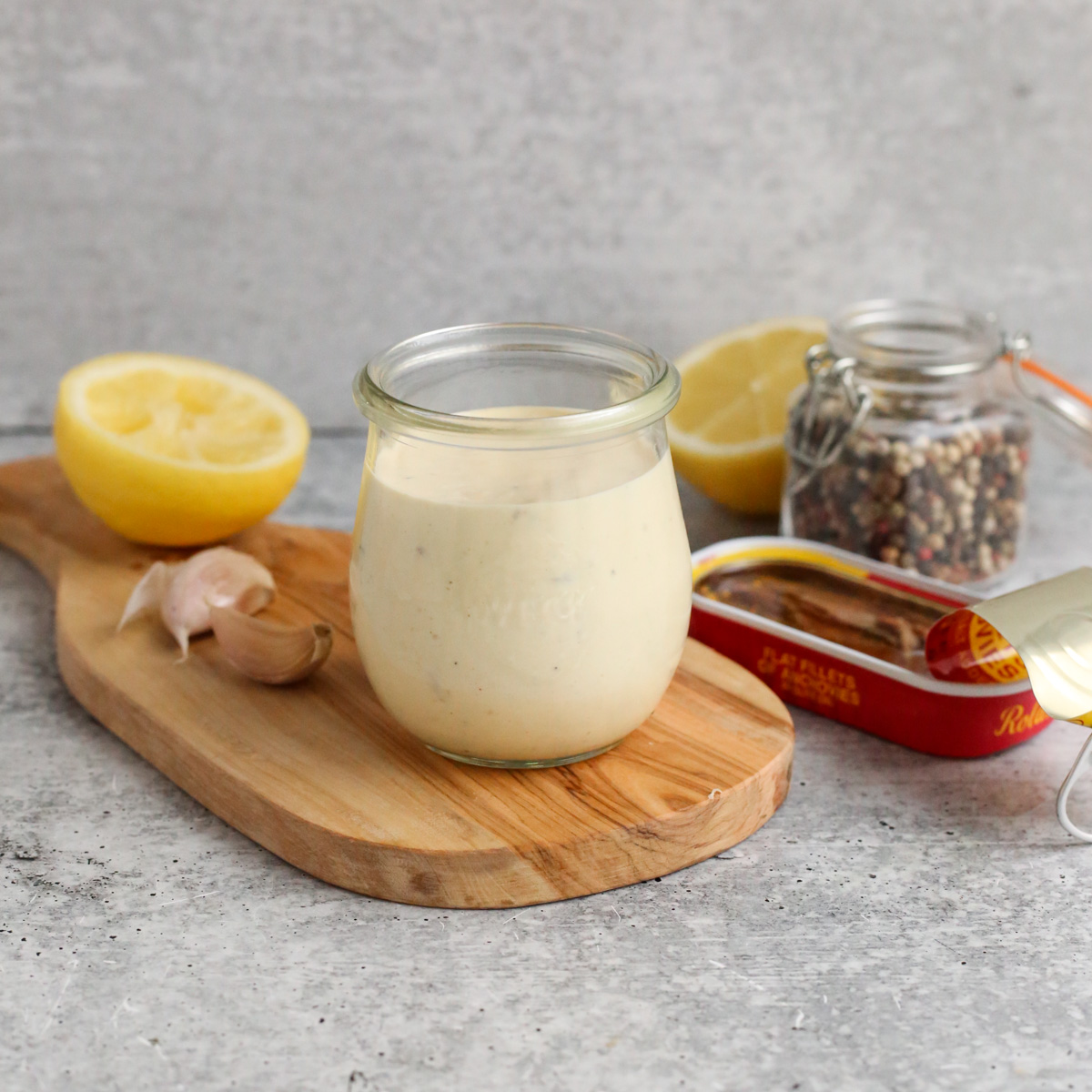 A clear glass jar filled to the top with a creamy homemade caesar salad dressing is displayed on a kitchen countertop with an open tin of anchovy fillets, a jar of peppercorns, garlic cloves, and a fresh lemon sliced in half