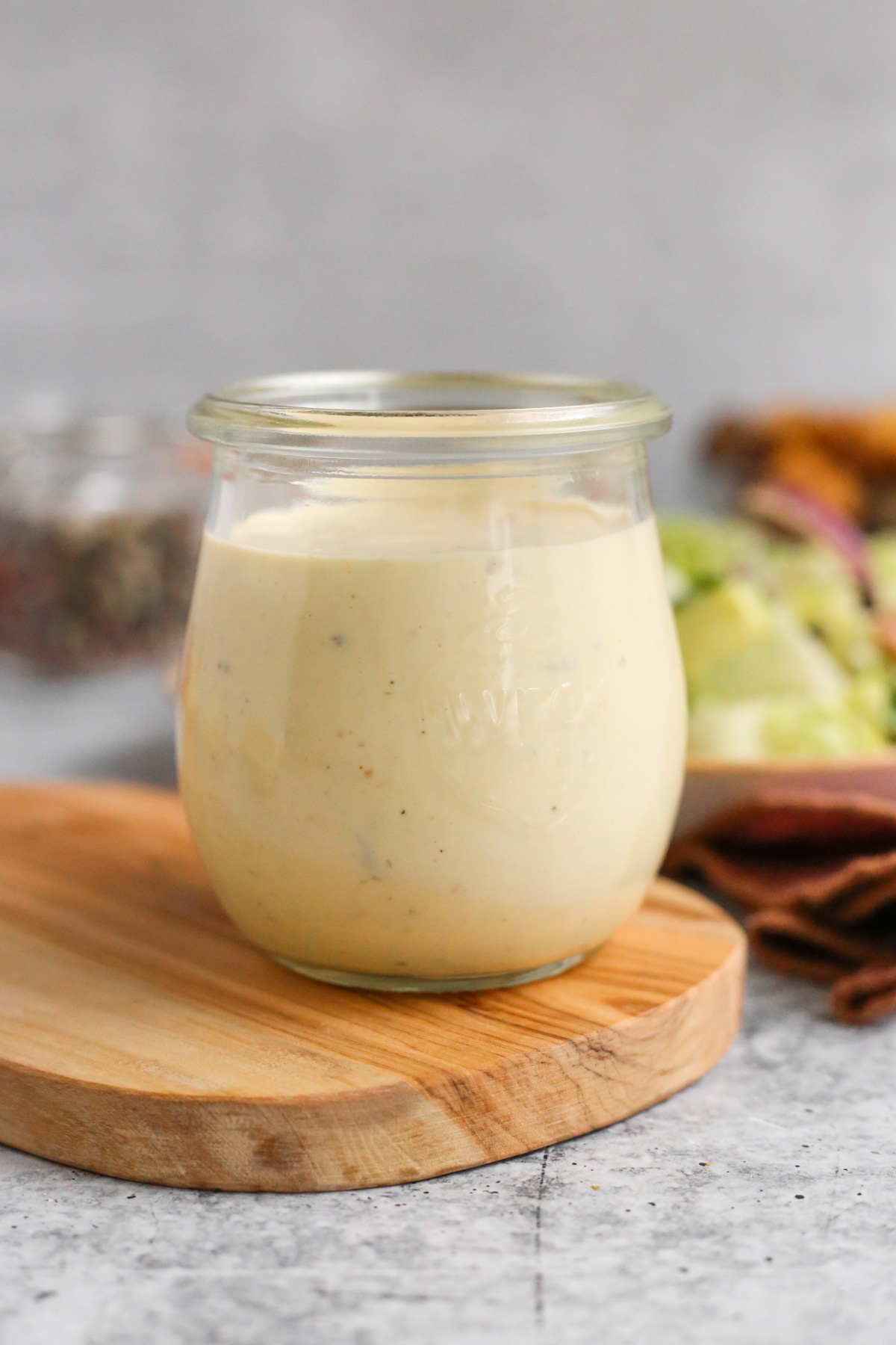 A small glass jar filled with a creamy homemade caesar salad dressing sits on a wooden serving board with a dark pink linen, plate of caesar salad, and jar of peppercorns visible in the background