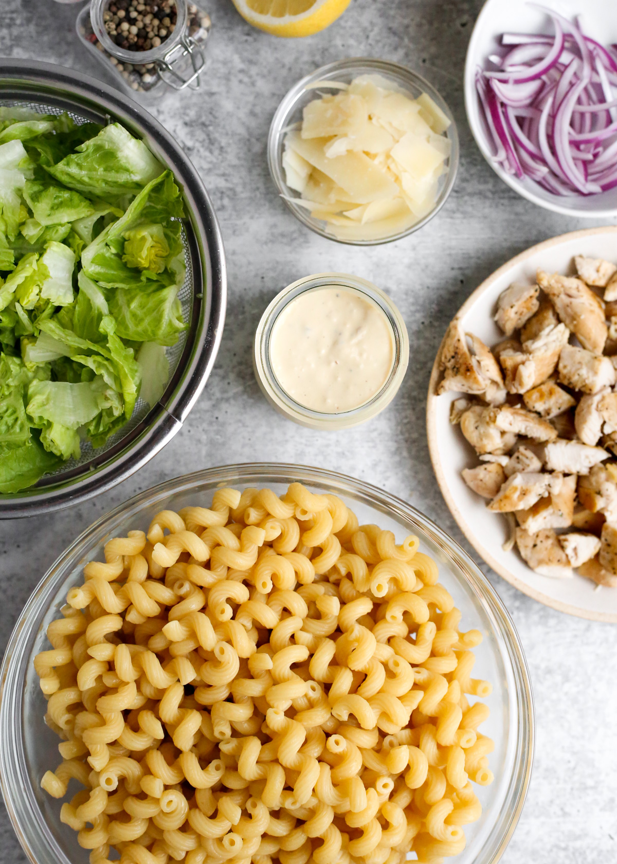 Flatlay style image of the ingredients needed to make a chicken caesar pasta salad, including crispy romaine lettuce, parmesan cheese, slide red onions, a creamy Caesar Salad dressing, seasoned chicken, and cooked pasta