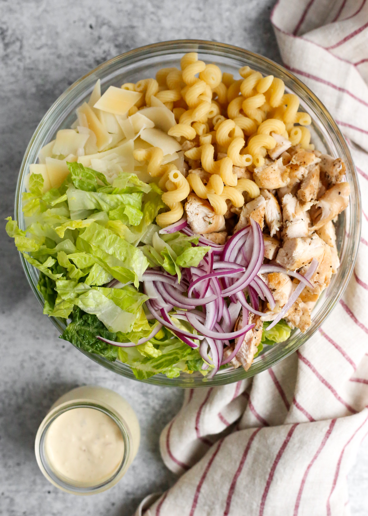 An overhead view of a creamy chicken Caesar pasta salad in a clear glass mixing bowl, before all ingredients are mixed together. The salad features cooked pasta, tender chicken pieces, crisp romaine lettuce, sliced red onions, and grated Parmesan cheese