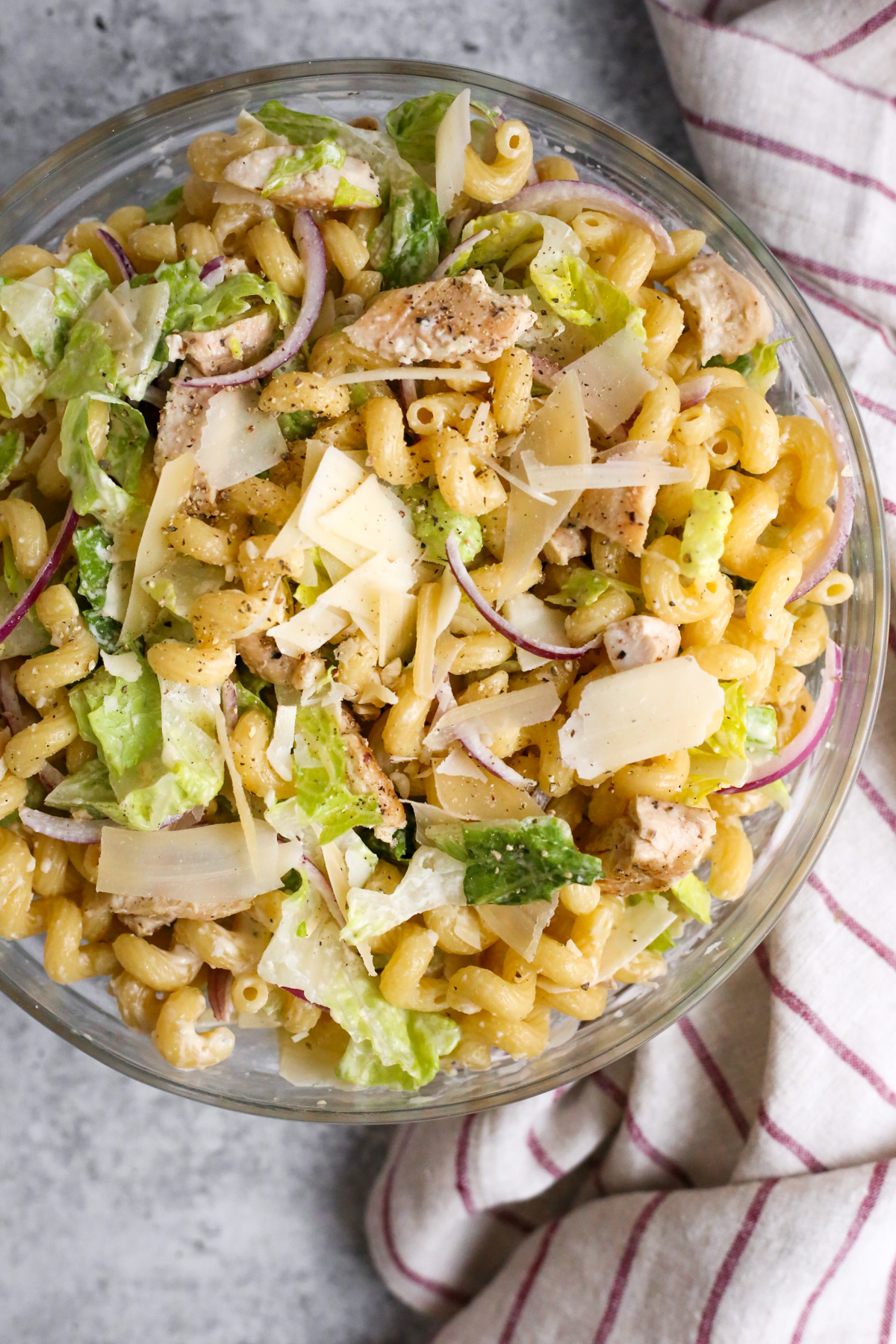 An overhead view of a creamy chicken Caesar pasta salad in a clear glass mixing bowl. The salad features cooked pasta, tender chicken pieces, crisp romaine lettuce, sliced red onions, and grated Parmesan cheese. The creamy Caesar dressing coats the ingredients, creating a delicious and visually appealing dish