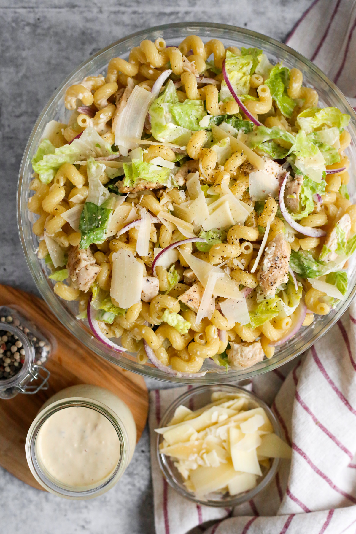An overhead view of a creamy chicken Caesar pasta salad in a clear glass mixing bowl. The salad features cooked pasta, tender chicken pieces, crisp romaine lettuce, sliced red onions, and grated Parmesan cheese. The creamy Caesar dressing coats the ingredients, creating a delicious and visually appealing dish