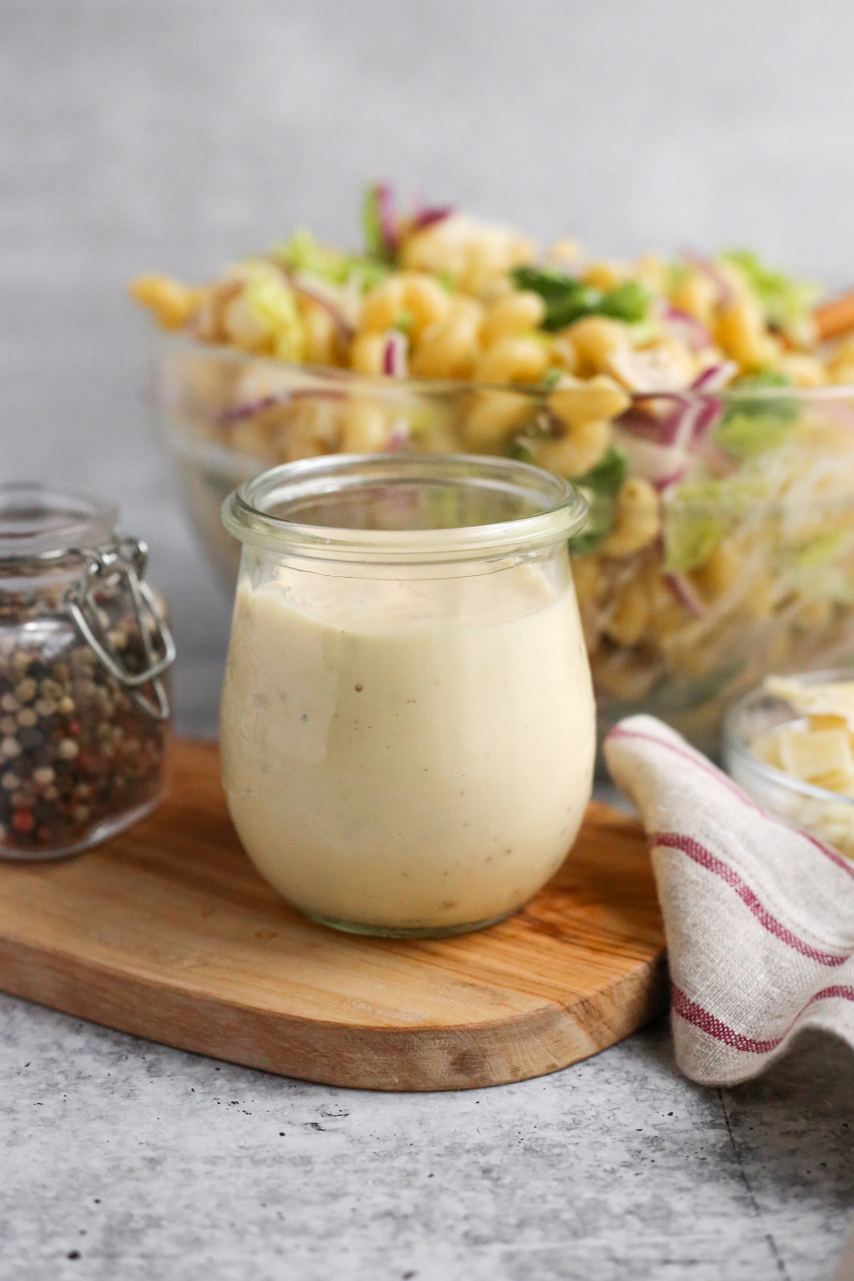 A clear glass jar full of creamy Caesar Salad dressing displayed on a small wooden serving platter. In the background, additional ingredients and a serving bowl full of a creamy pasta salad is visible