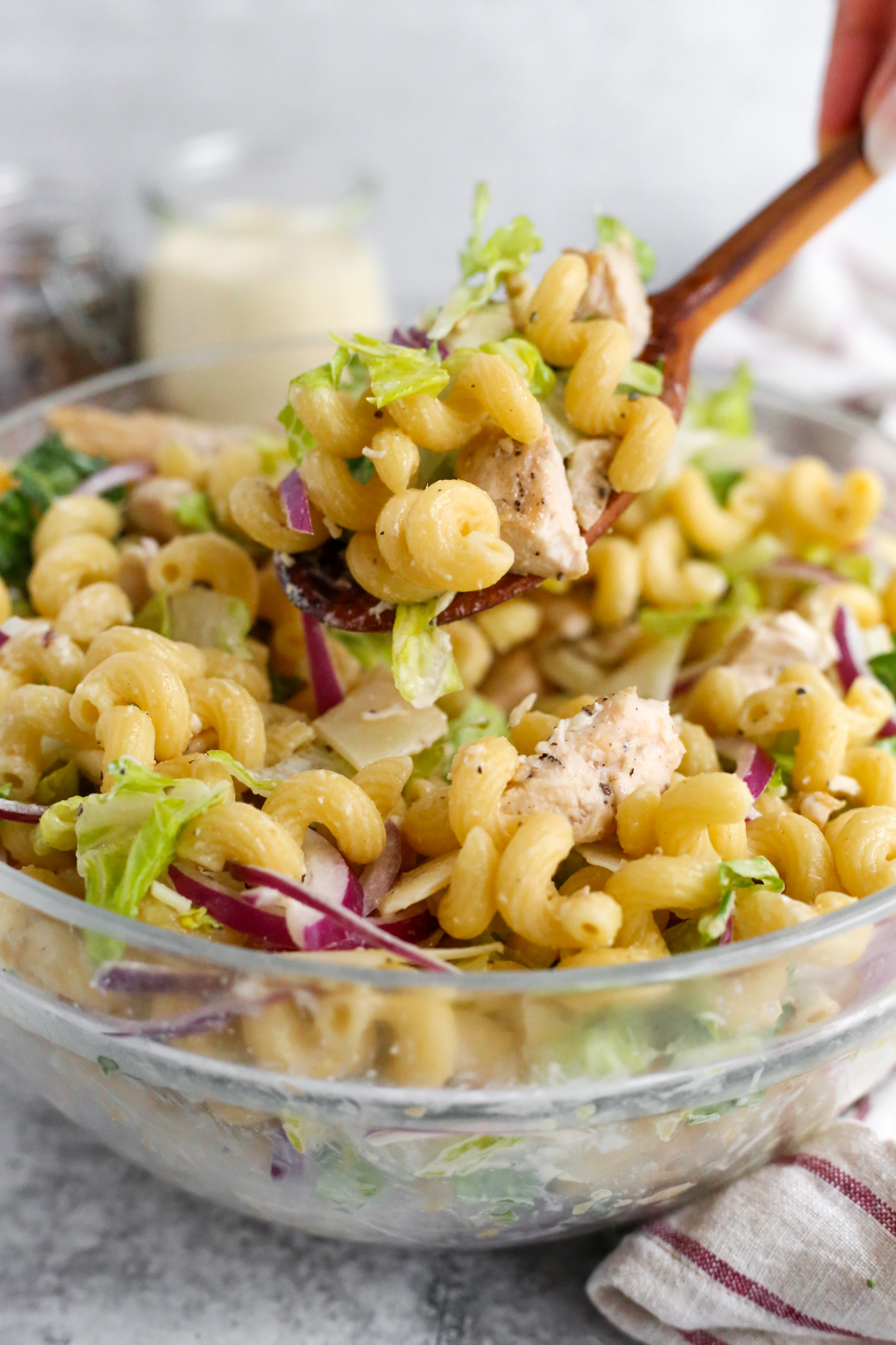 A woman's hand holds a wooden serving spoon piled high with creamy chicken caesar pasta salad. The pasta is cooked al dente and the seasoned chicken looks juicy and tender. The crispy romaine lettuce and sliced red onions are mixed evenly throughout the pasta salad