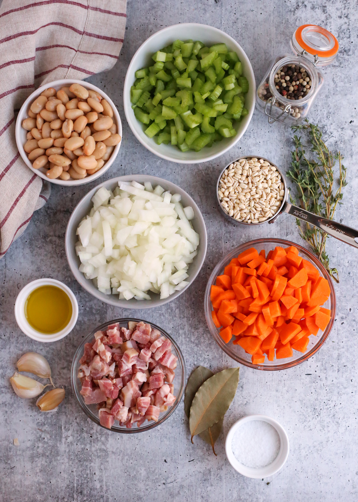 Assortment of ingredients to make a bean and barley soup recipe, including canned white beans, uncooked barley in a measuring cup, diced onions, carrots, and celery, bay leaves, uncooked pancetta, garlic cloves, a small ramekin of olive oil, and sprigs of fresh thyme 