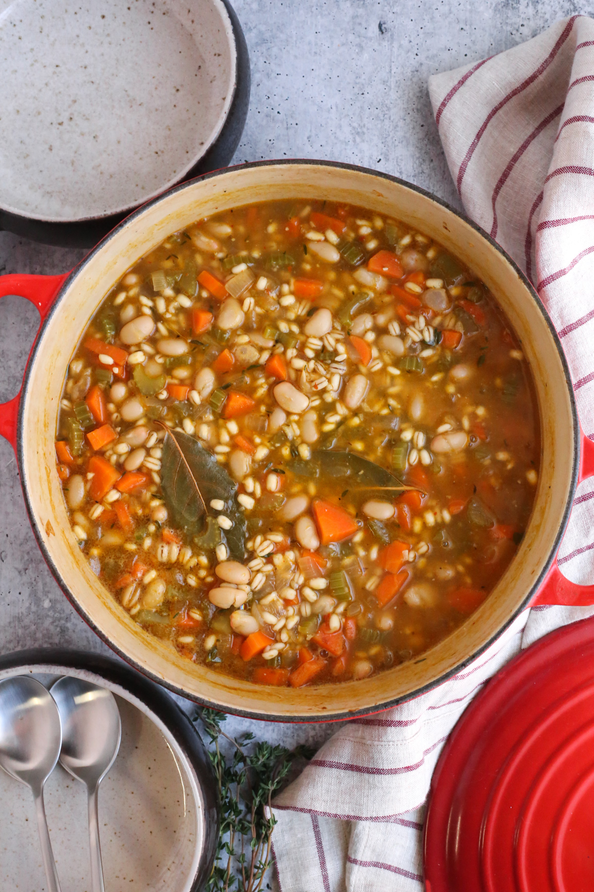 Overhead view into a Dutch oven used to cooked a bean and barley soup recipe. The soup is partially cooked with bay leaves and other ingredients visible in the broth