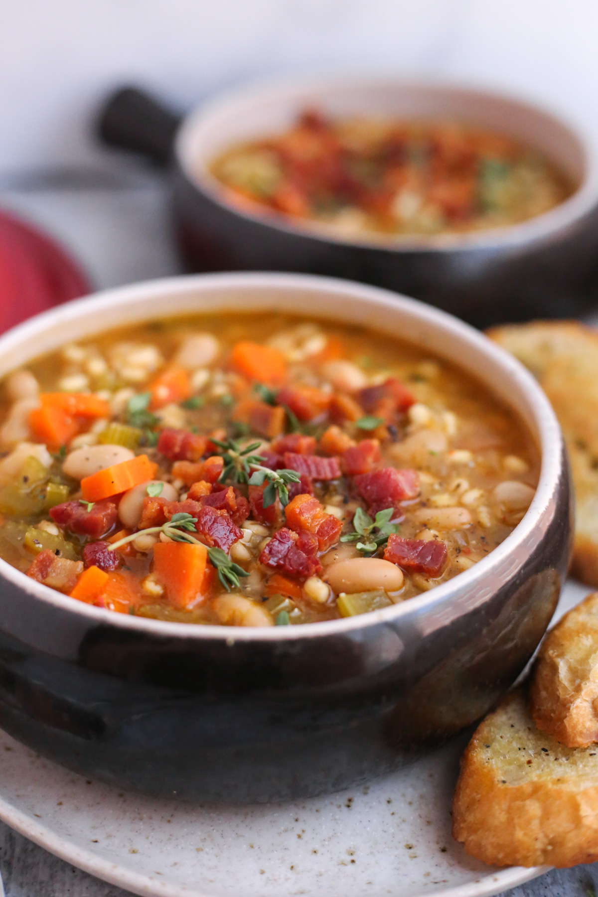 A close angled view into a soup bowl that holds a full serving of Great Northern Bean Soup. The soup looks savory and filling, topped with pancetta pieces and fresh thyme
