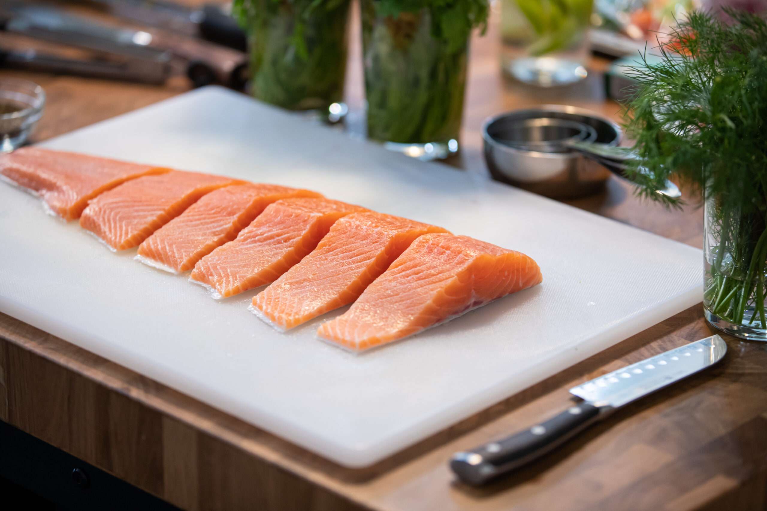 A kitchen scene with a cutting board displaying a large fillet of fresh salmon, arranged with a chefs knife, measuring cups, cooking utensils, and fresh herbs