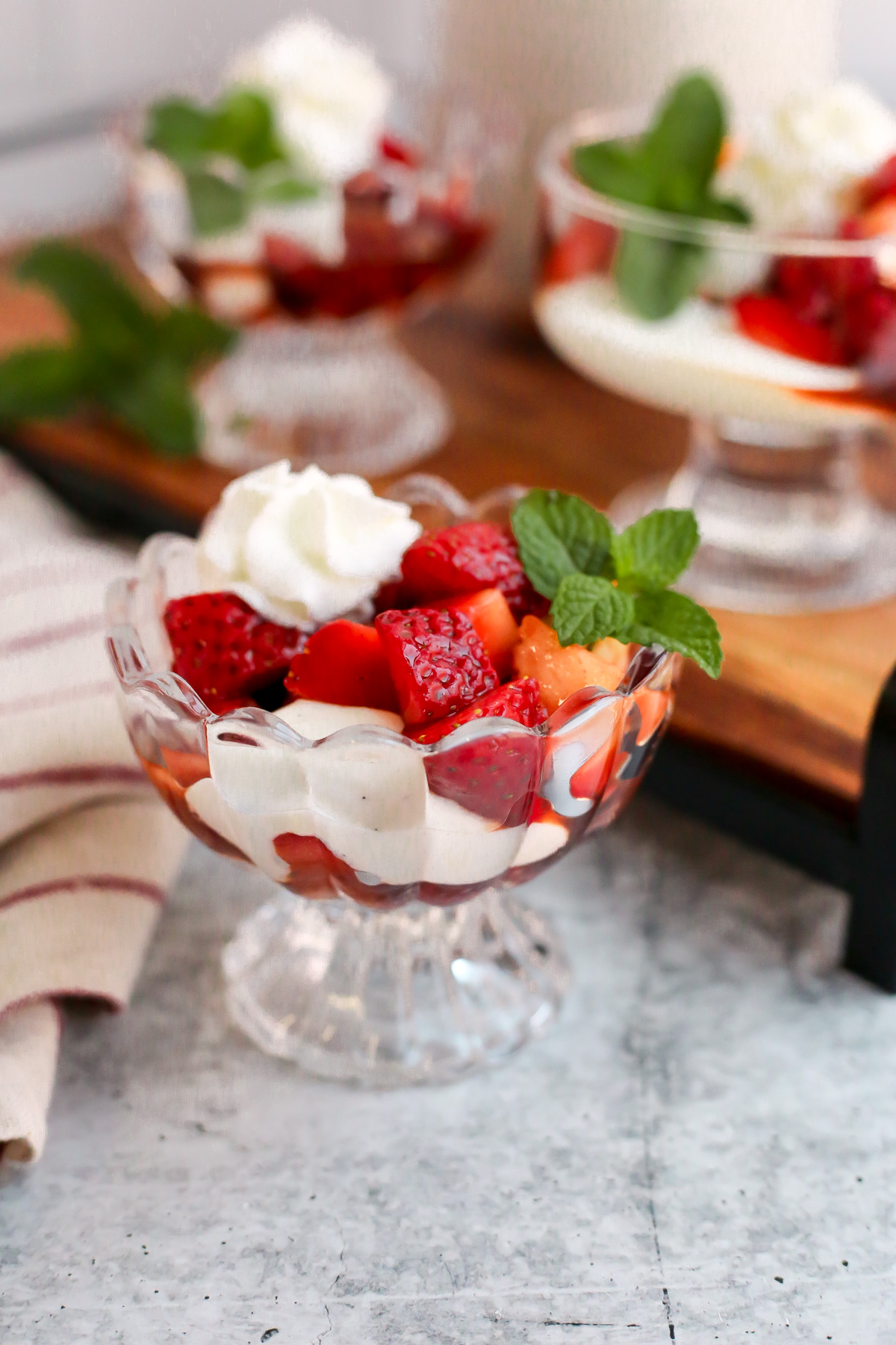 Angled view of a clear glass serving dish filled with Balsamic Macerated Strawberries and Cream. A fresh sprig of mint decorates the side of the dish