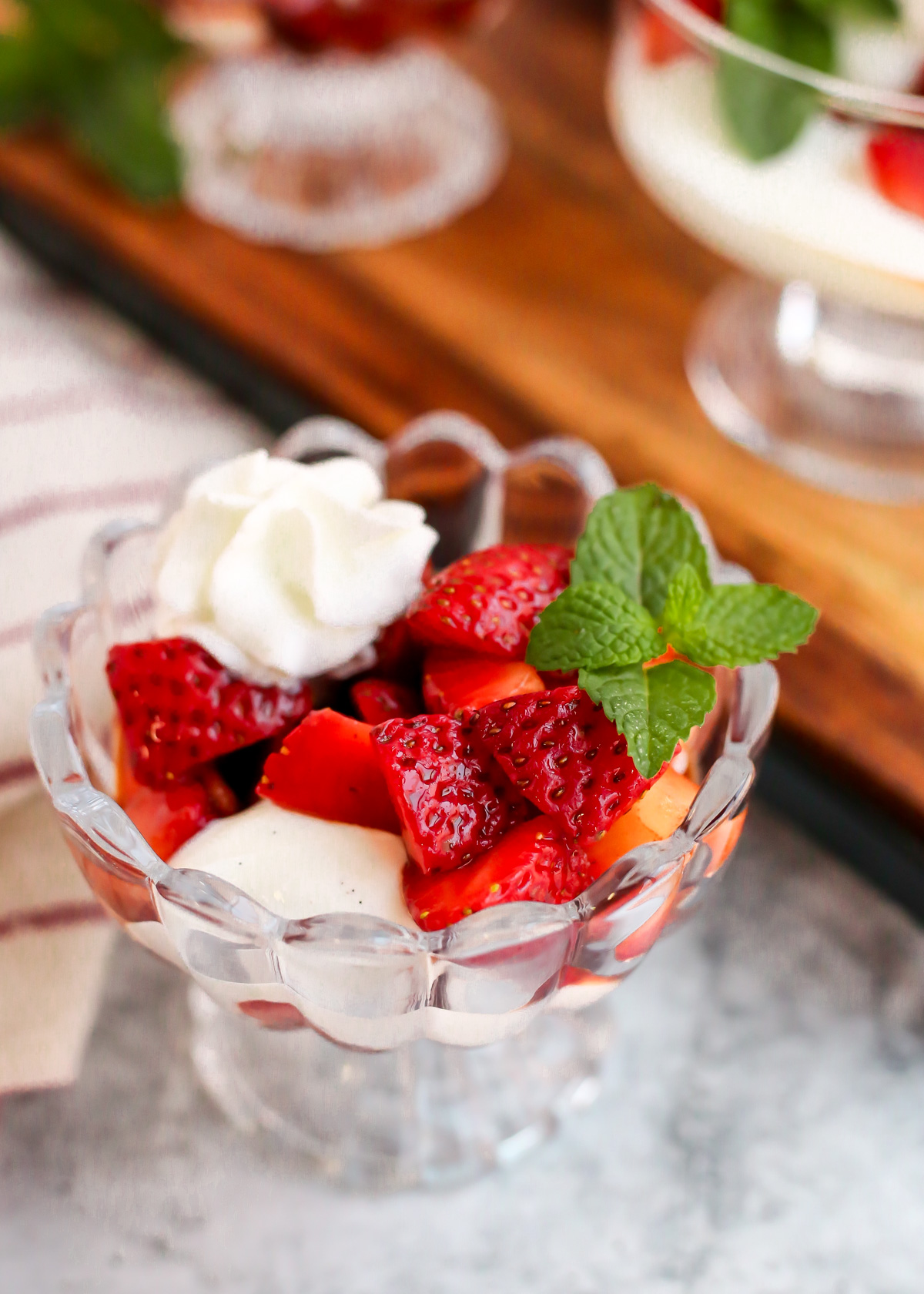 Angled view of a glass serving dish filled with balsamic macerated strawberries and cream, garnished with a sprig of fresh mint leaves and some fresh whipped cream