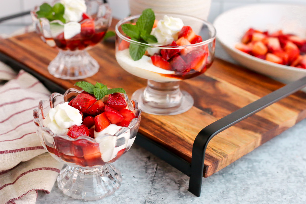 Glass serving dishes filled with balsamic macerated strawberries and cream displayed on a wooden serving platter on a kitchen counter, landscape orientation