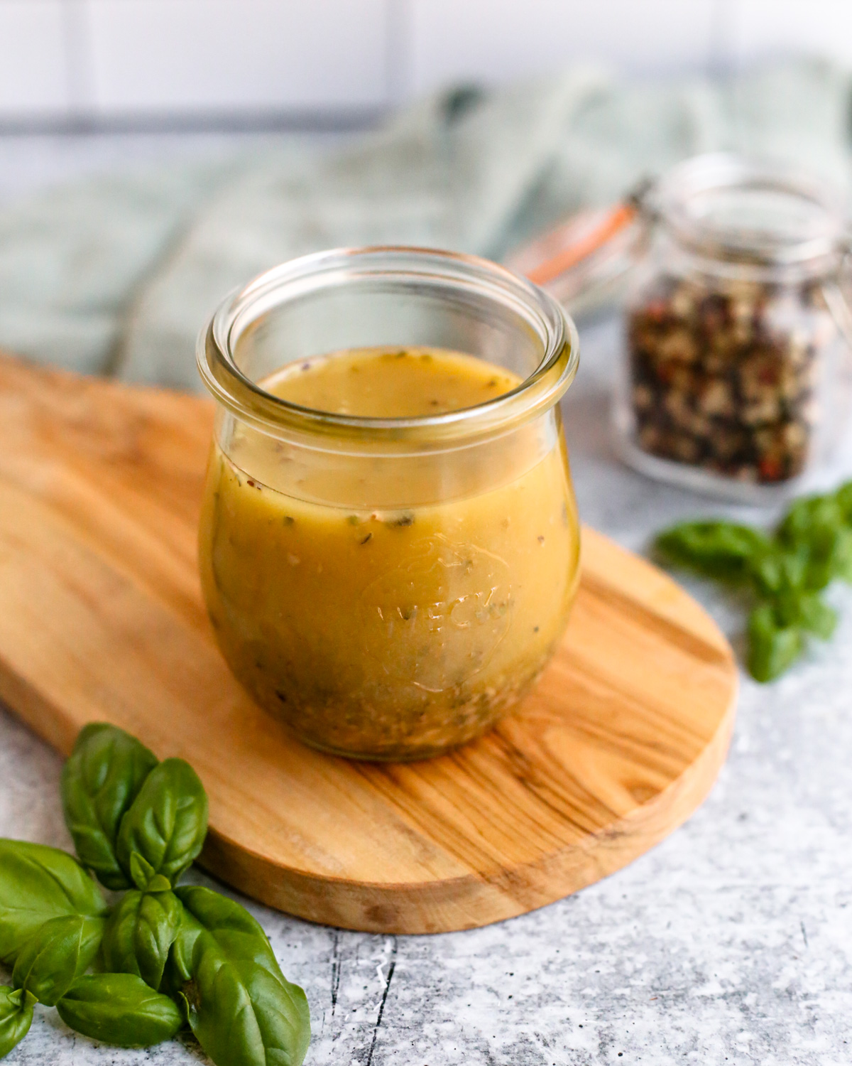 A small glass jar filled to the top with a homemade red wine vinaigrette salad dressing is displayed on a small wooden serving board on a kitchen countertop, surrounded by peppercorns and fresh basil