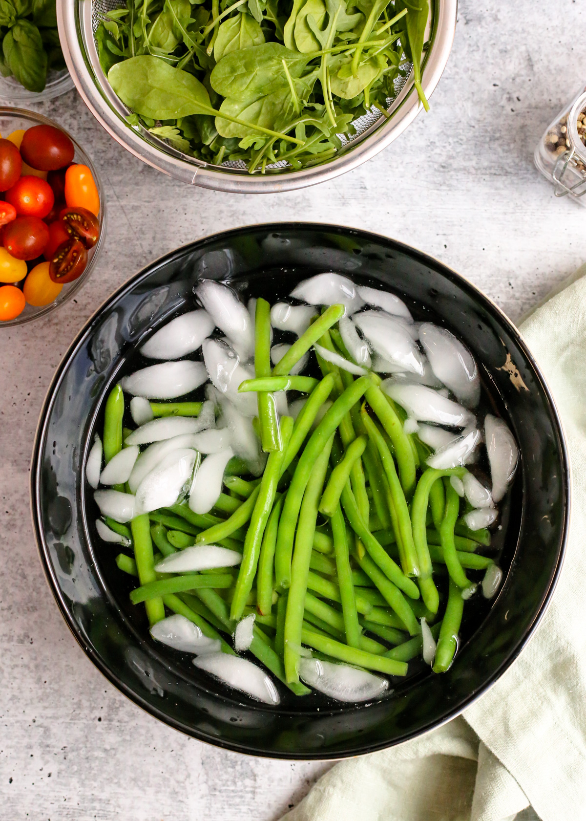 Overhead view of a black ceramic bowl filled with blanched green beans surrounded by water and ice cubes. The green beans are partially cooked but vibrant green and fresh looking