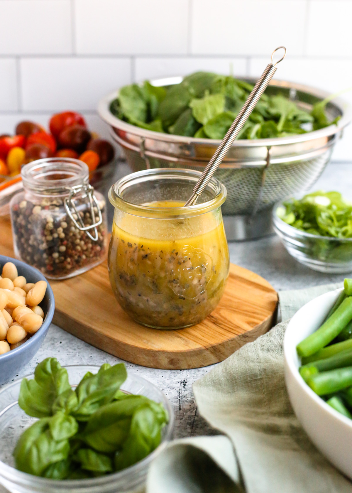 A small glass jar filled with a homemade red wine vinaigrette is sitting on small wooden display board and surrounded by various colorful fresh vegetables as if in preparation to make a delicious, fresh salad